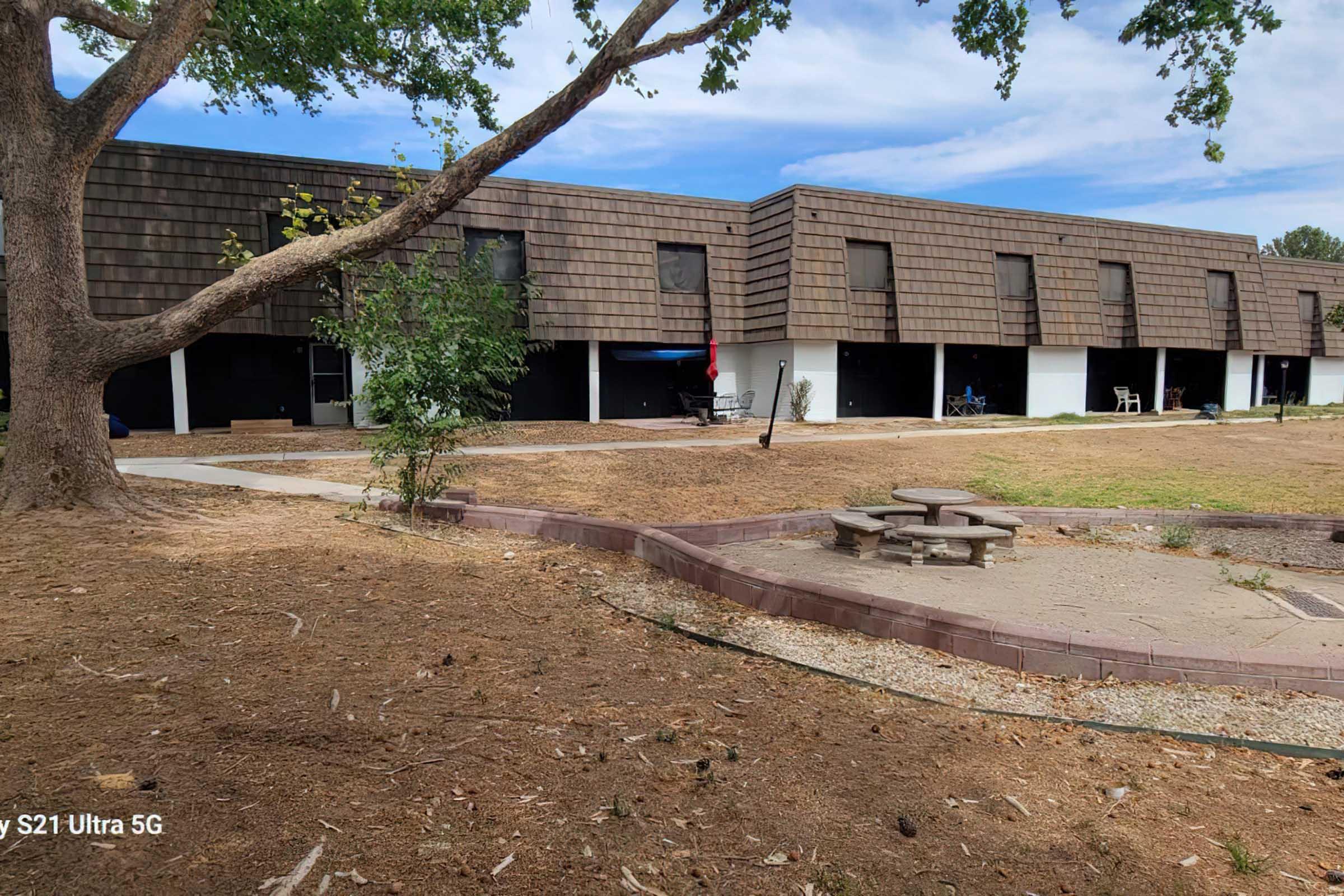 A view of a residential building with a brown shingle exterior. In the foreground, a circular stone fire pit is surrounded by a small grassy area. There is a large tree to the left with green leaves, and the ground is mostly dry with some scattered patches of grass. The sky is clear and bright.