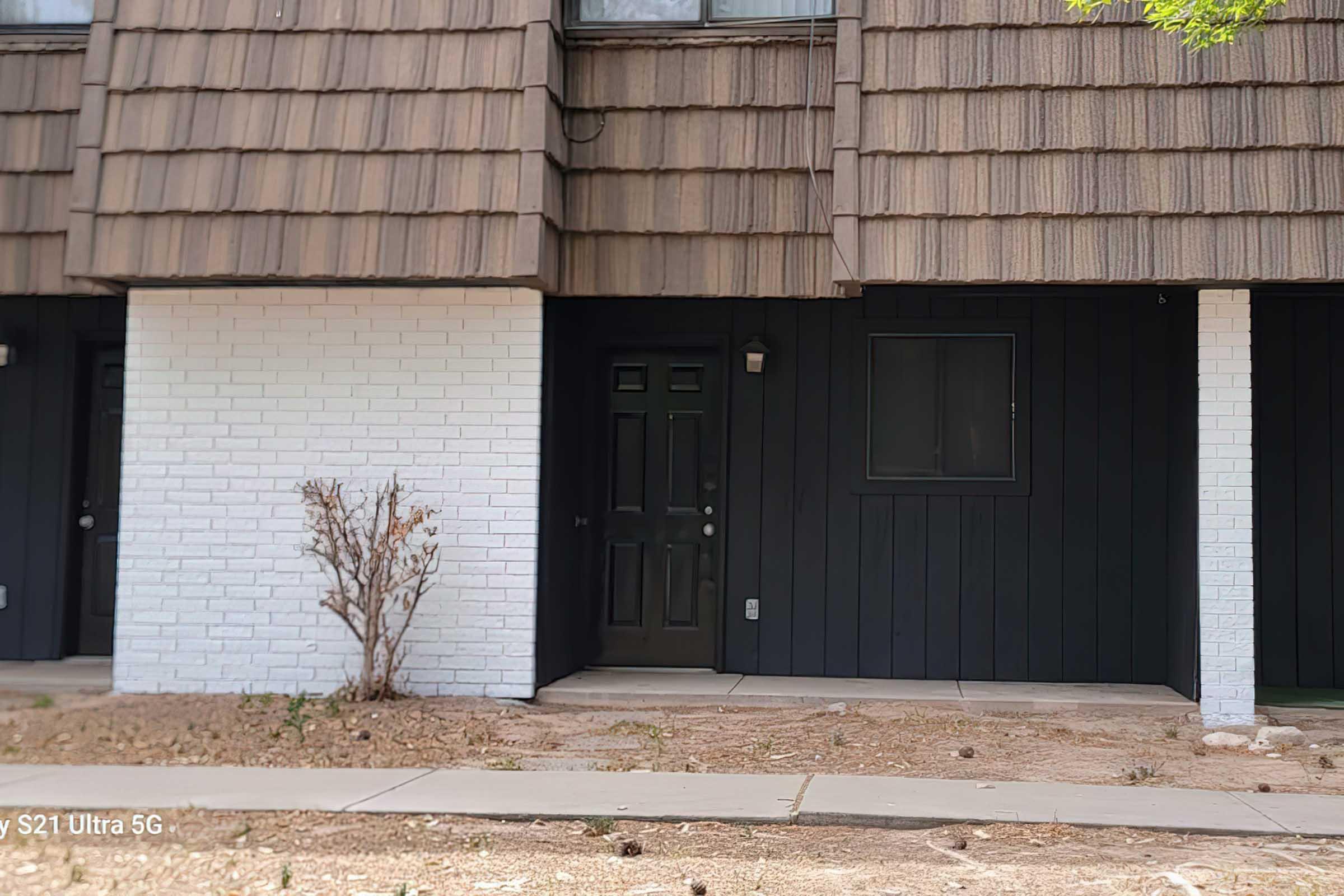 A close-up view of a building entrance featuring a dark green door, white brick and dark wood siding. The area is landscaped with minimal plants, and the ground is mostly bare soil. A small window is visible next to the door. There are wooden shingles on the upper part of the building.