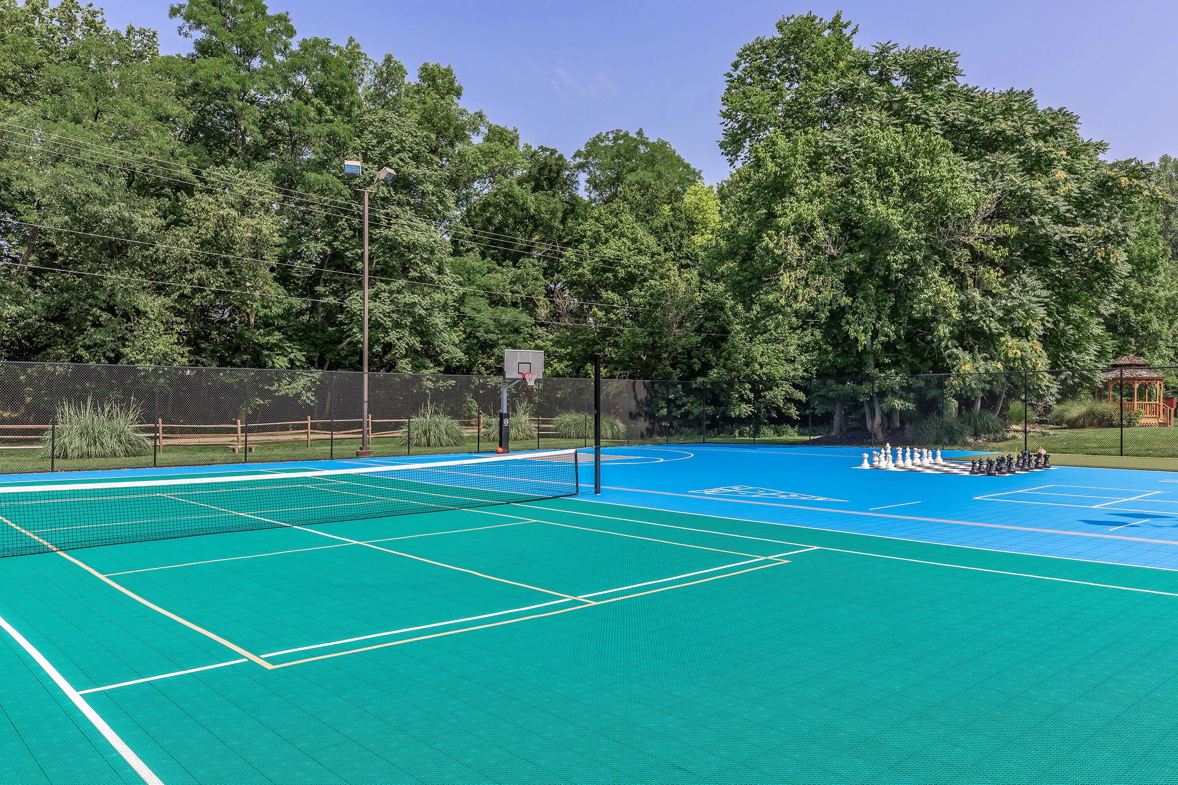 A vibrant sports court area featuring a blue and green tennis court with a net, surrounded by lush trees. In the background, there's a basketball hoop and a section for other games with equipment neatly arranged. The bright blue sky complements the lively outdoor setting.