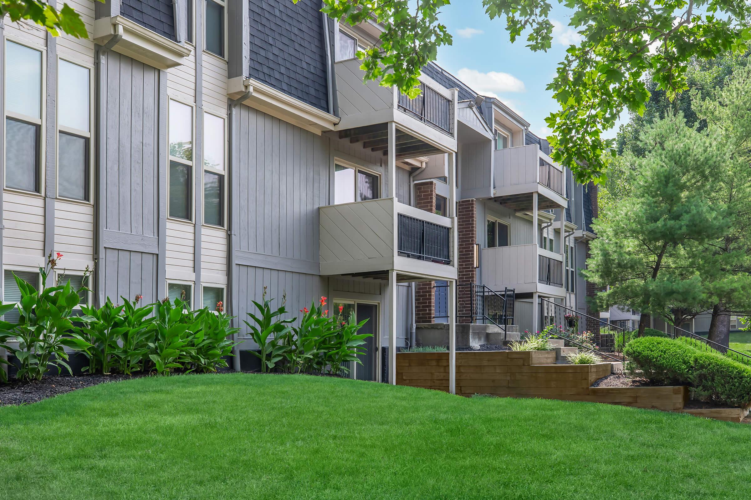 View of a modern apartment community featuring multiple gray and beige buildings with balconies. Lush green grass and landscaping with vibrant plants in the foreground. Trees are visible in the background under a clear blue sky, creating a serene residential atmosphere.