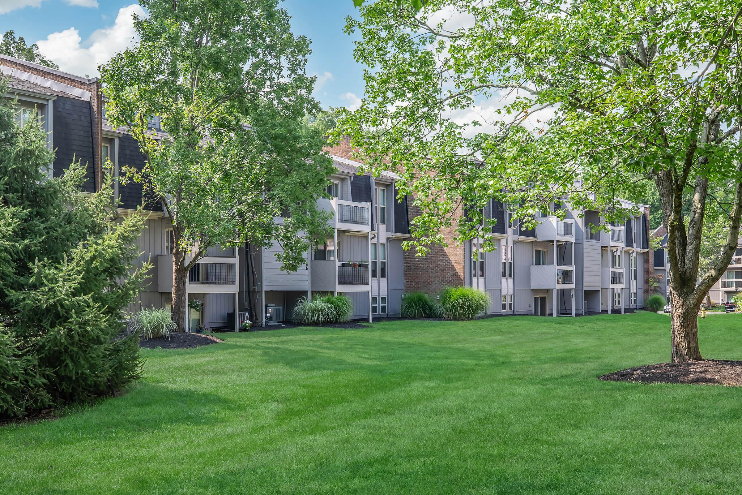 A residential apartment community featuring multiple units, surrounded by lush green grass and trees. The buildings have balconies and are designed with a mix of brick and siding. The scene includes clear blue skies and a well-maintained outdoor area.