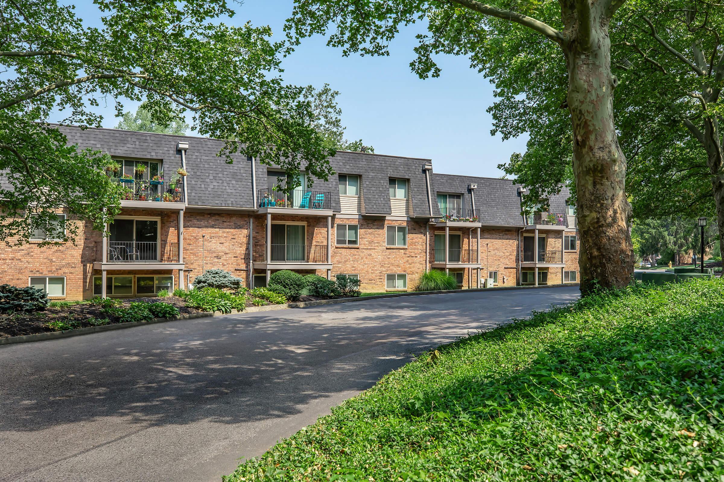 A row of brick apartment buildings with multiple balconies surrounded by green trees and shrubs. A paved street runs alongside the buildings, and colorful patio furniture is visible on the balconies. The scene is sunny, showcasing a peaceful residential area.