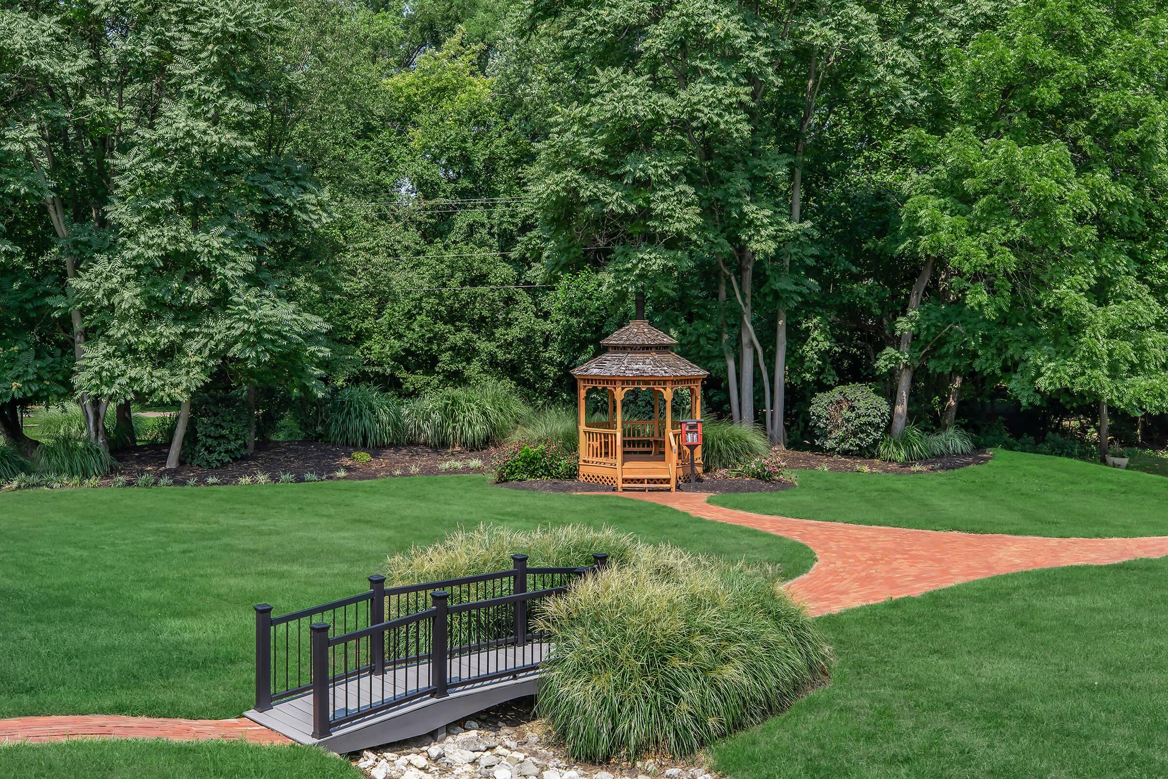 A lush green garden featuring a wooden gazebo surrounded by trees. There is a red brick pathway leading to the gazebo, and a small black bridge crossing over a rocky area. The scene is well-maintained, showcasing vibrant greenery and a tranquil atmosphere.