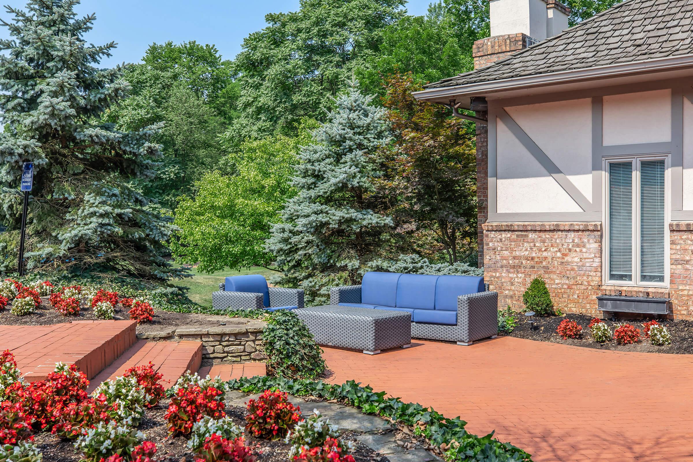 A cozy outdoor seating area featuring a blue sofa set surrounded by vibrant red and green flowers. The patio is made of brick, leading to a well-maintained lawn and lush trees in the background, creating a serene and inviting atmosphere. A stone border adds a rustic touch to the landscape.