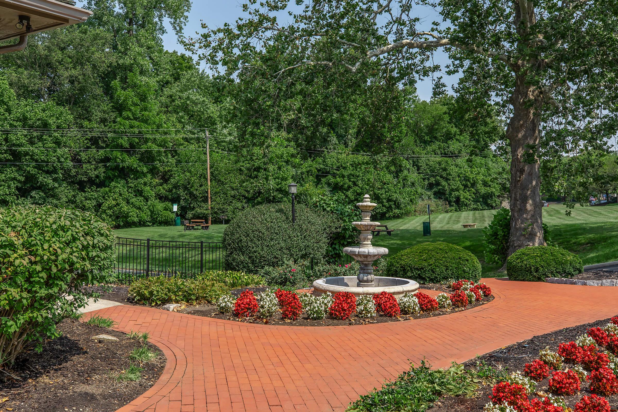 A beautifully landscaped garden featuring a stone fountain at the center, surrounded by vibrant red flowers and neatly trimmed bushes. The pathway is made of red bricks, leading towards a green lawn and trees in the background, creating a serene outdoor setting.