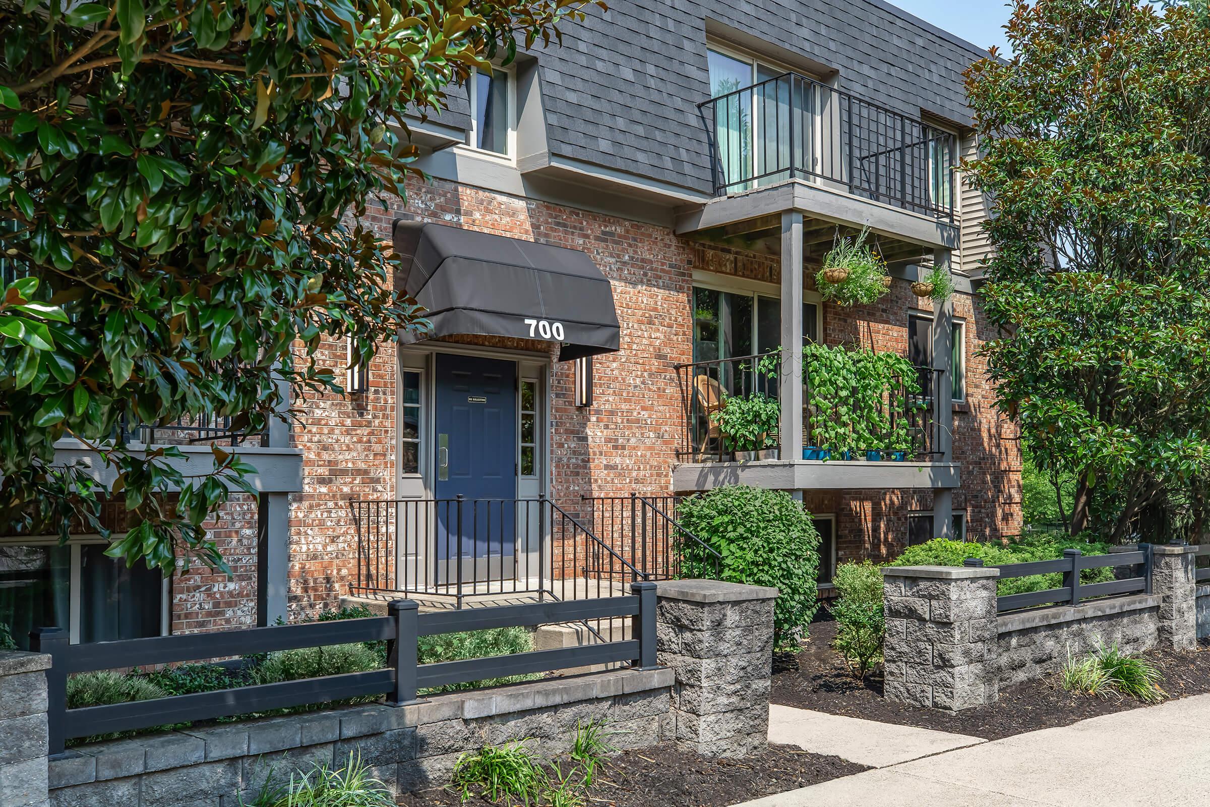 A modern brick multi-story building with balconies and greenery, featuring a dark awning over the entrance. The address "700" is displayed on the door, surrounded by well-maintained landscaping, including shrubs and plants. A concrete pathway leads to the entrance.