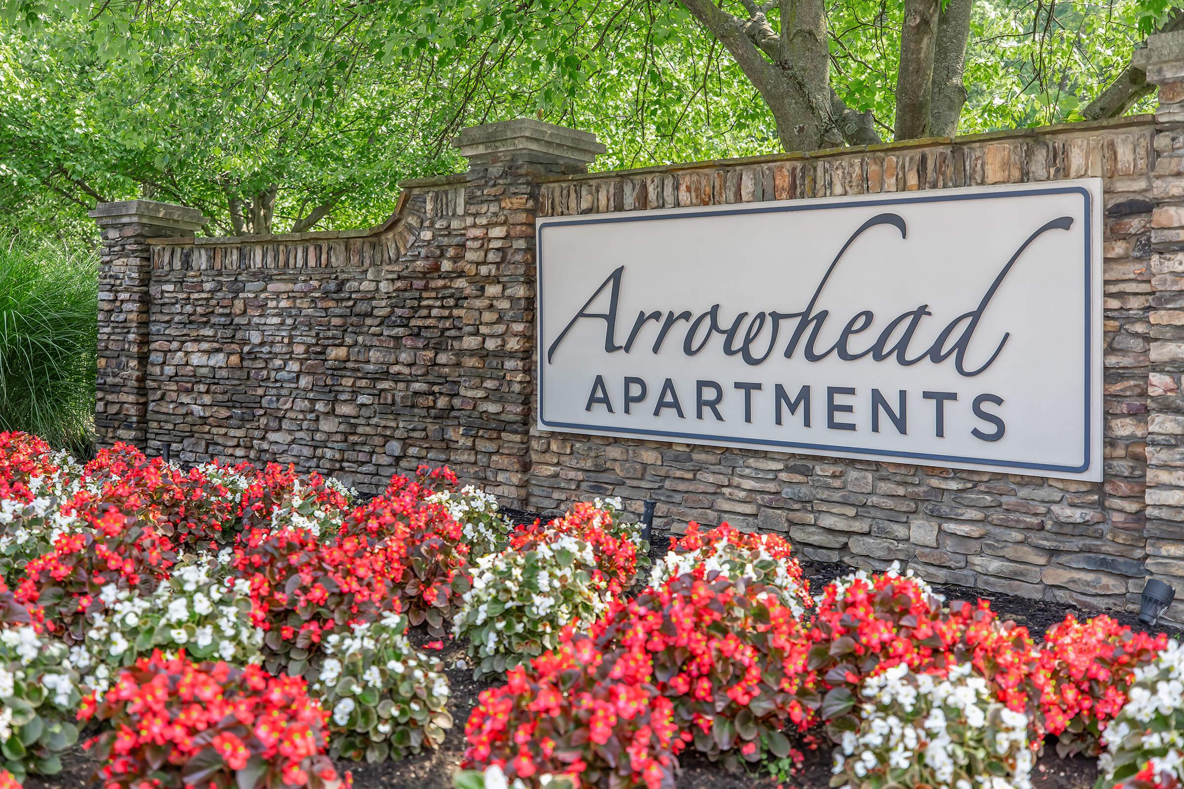 A stone wall featuring a sign that reads "Arrowhead Apartments" in elegant script. Colorful red and white flowers are planted at the base, surrounded by lush green foliage, creating a welcoming entrance to the apartment community.