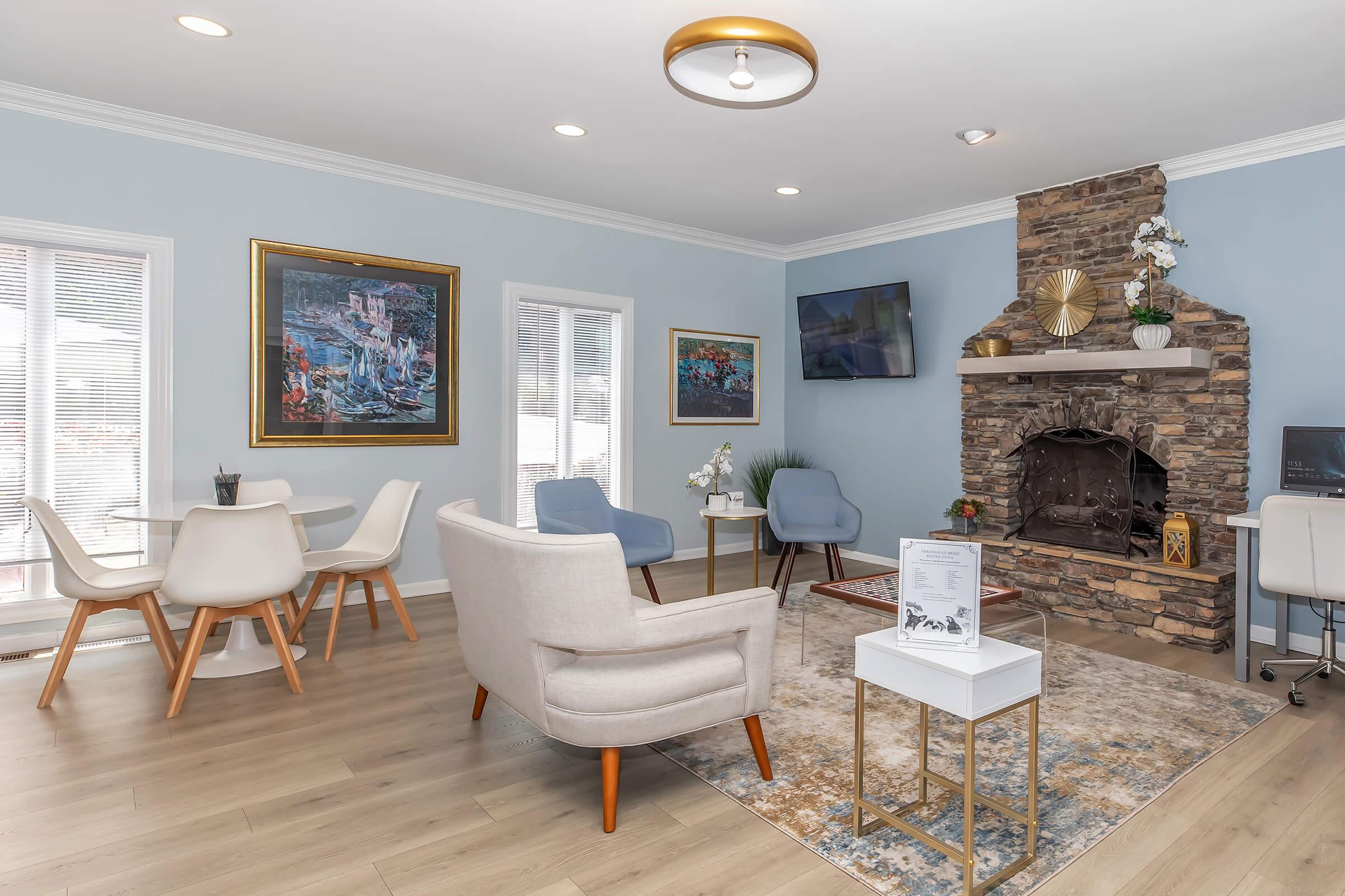 A cozy sitting area featuring light blue walls, modern chairs, and a stone fireplace. There are a few paintings on the walls, a round light fixture, and a decorative rug on the wooden floor. A small table is set up with a plant and information display, creating a welcoming atmosphere.