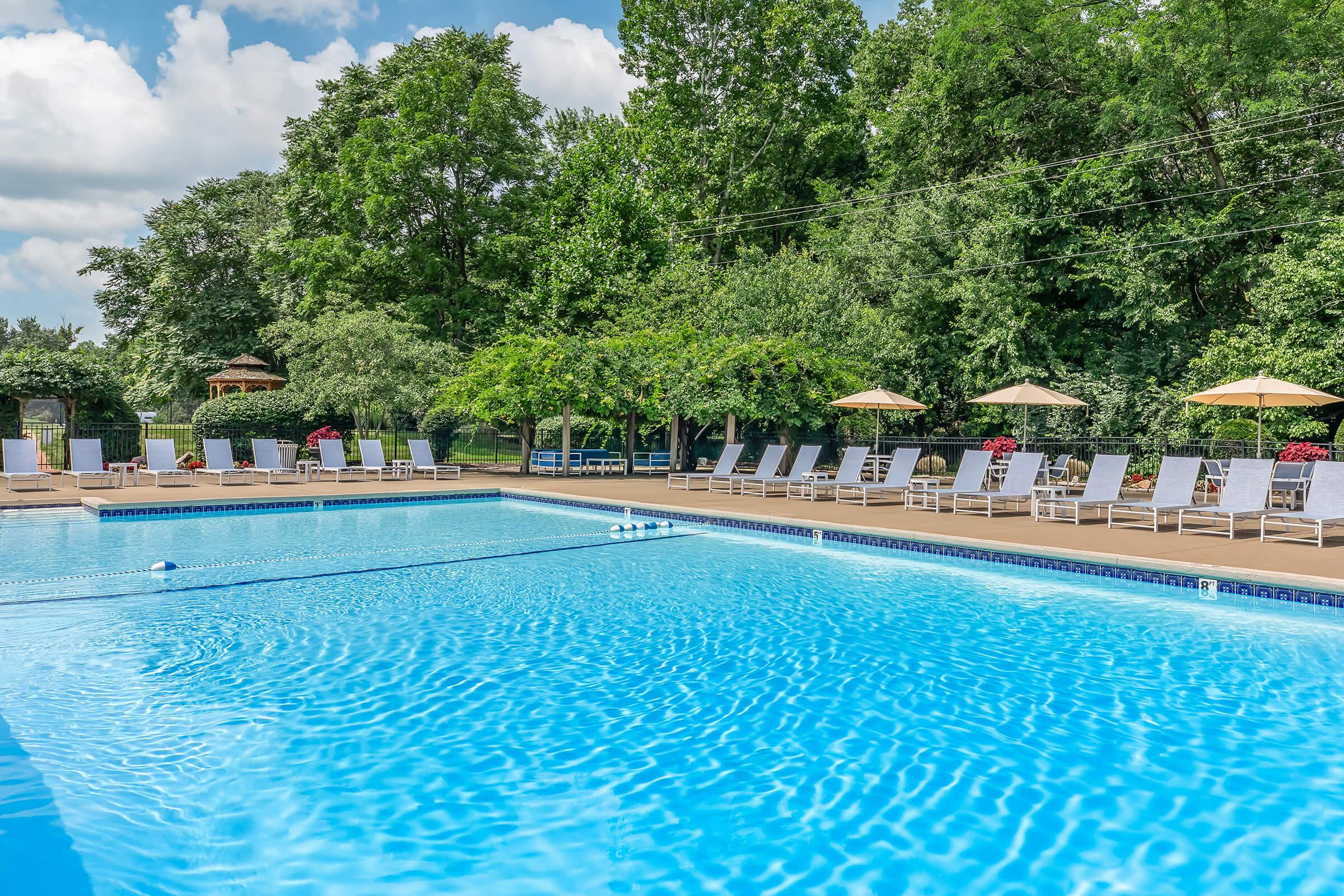 A clear blue swimming pool surrounded by lush greenery, lounge chairs, and umbrellas. The sun shines down on the water, creating a sparkling effect. In the background, there are trees and blooming flowers, adding to the serene atmosphere of the pool area.