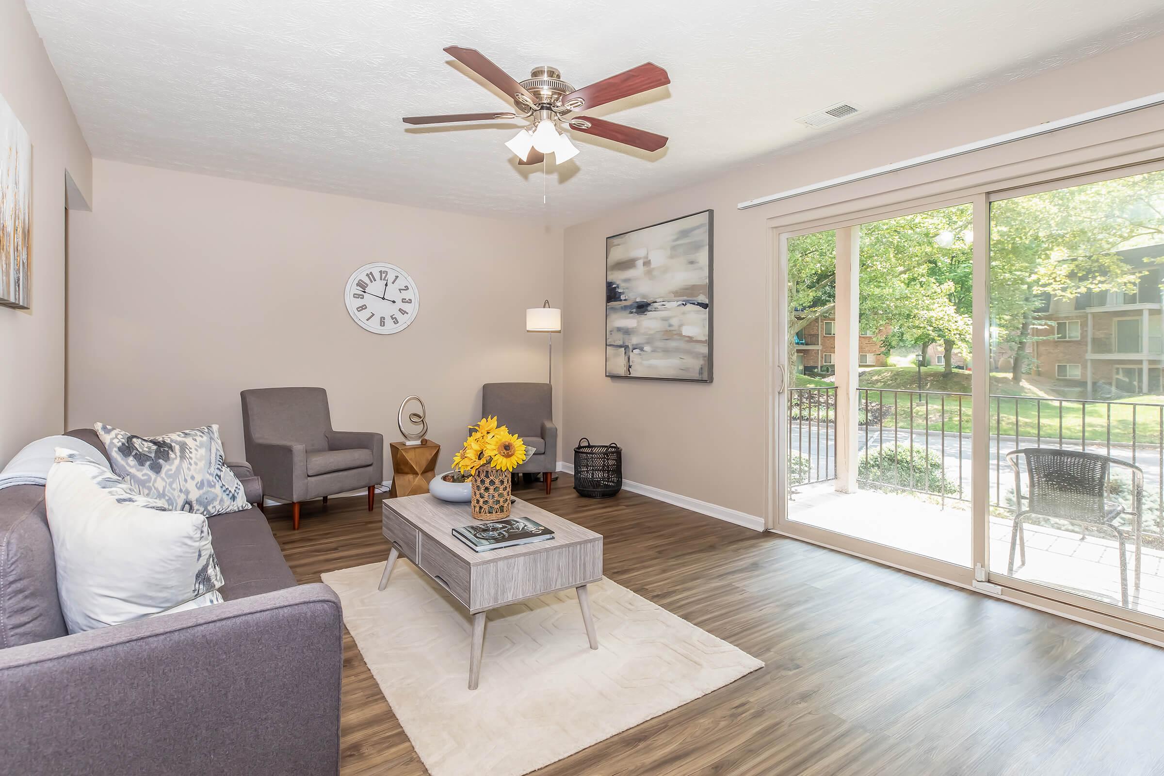 A modern living room featuring a gray sofa and two gray armchairs. A round wall clock is mounted above the sofa. A coffee table with books and a vase of yellow flowers sits in the center. Large sliding glass doors lead to an outdoor view. The flooring is light wood, and the walls are painted a soft beige.