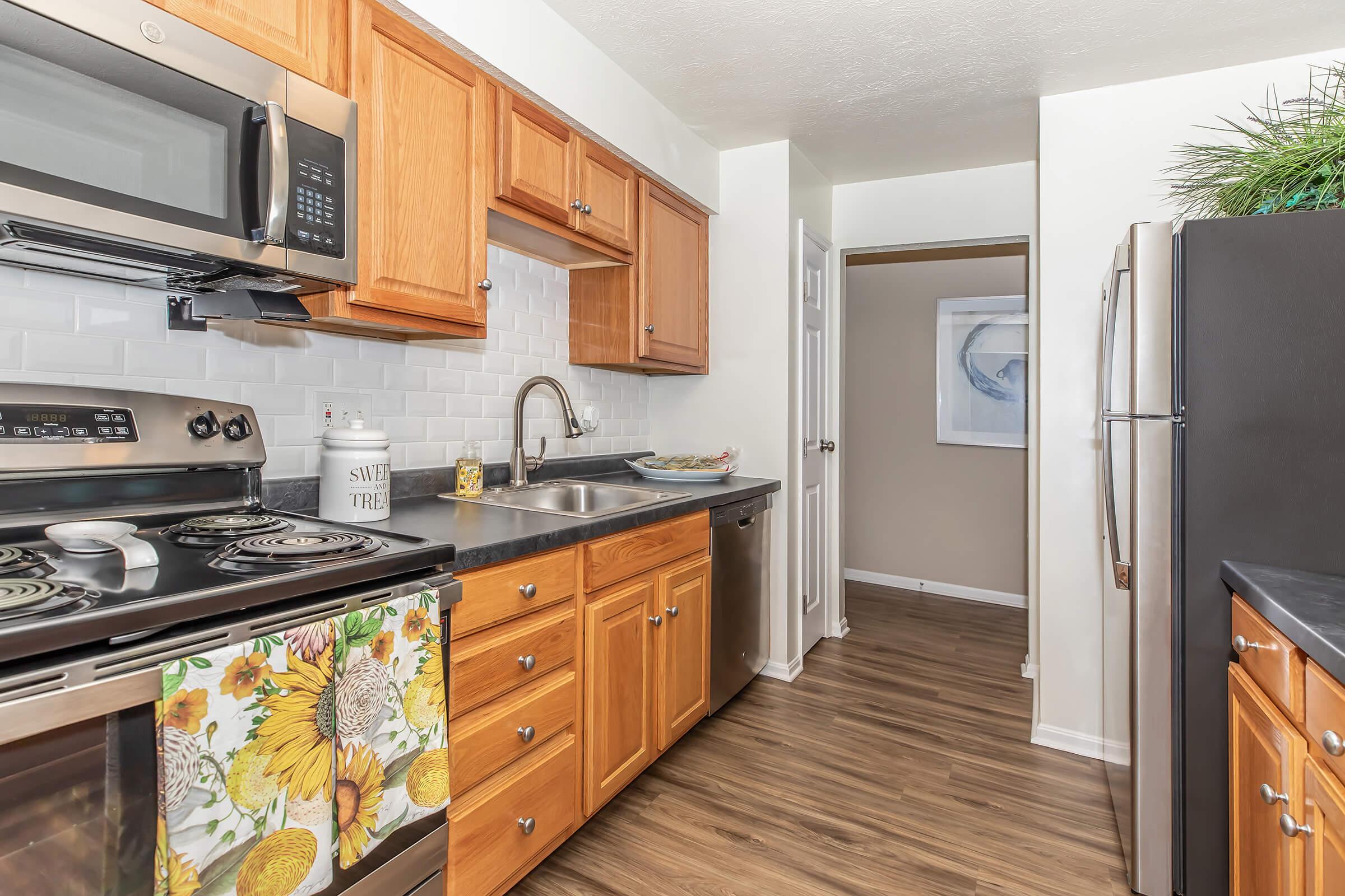 Modern kitchen featuring wooden cabinets, stainless steel appliances including a microwave and dishwasher, a double sink, and a tiled backsplash. The floor has a wood-like finish, and there is a small hallway leading to another room. Decor includes a floral kitchen towel and a decorative wall art piece.