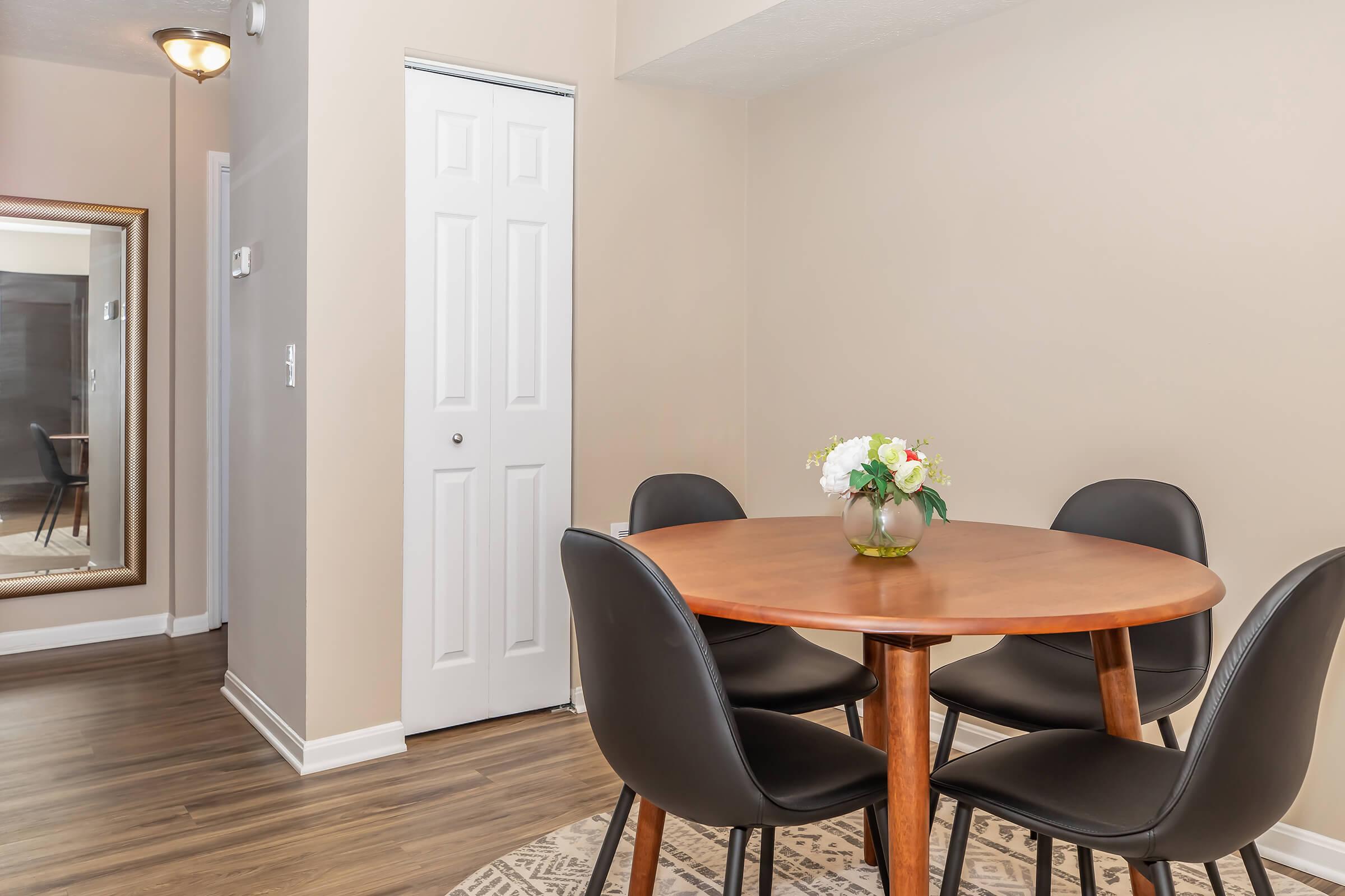 A cozy dining area featuring a round wooden table surrounded by four black chairs. A small vase with flowers sits on the table. The wall is painted in a warm tone, and there is a mirrored wall on the left, along with a white closet door. The floor has a wooden texture, enhancing the inviting atmosphere.