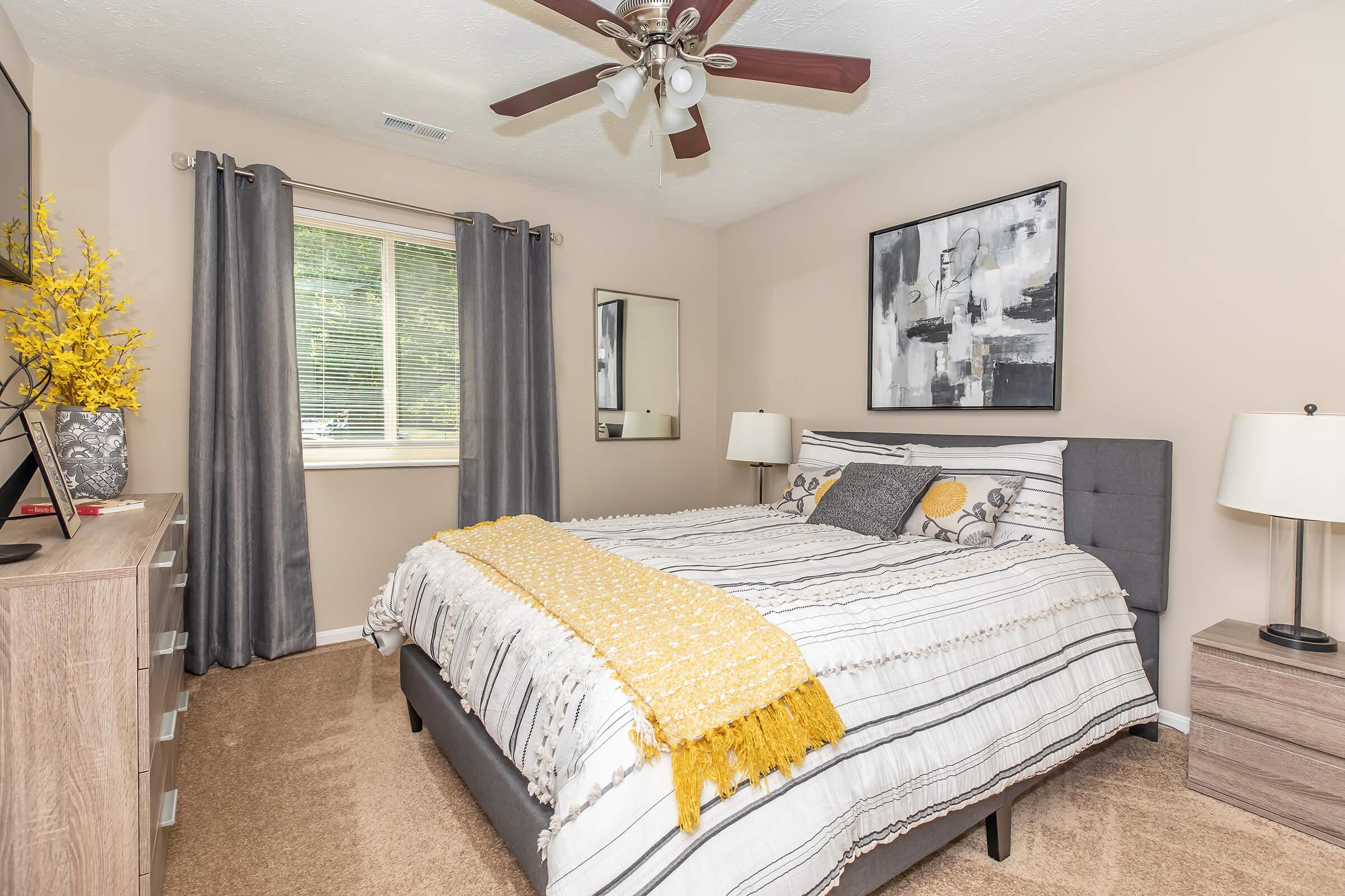 A cozy bedroom featuring a queen-sized bed with gray and white striped bedding, a mustard yellow throw blanket, and decorative pillows. There's a wooden dresser, a mirror, a TV on top, and large windows with sheer curtains. The walls are painted a soft beige, and there are decorative touches like a yellow plant.