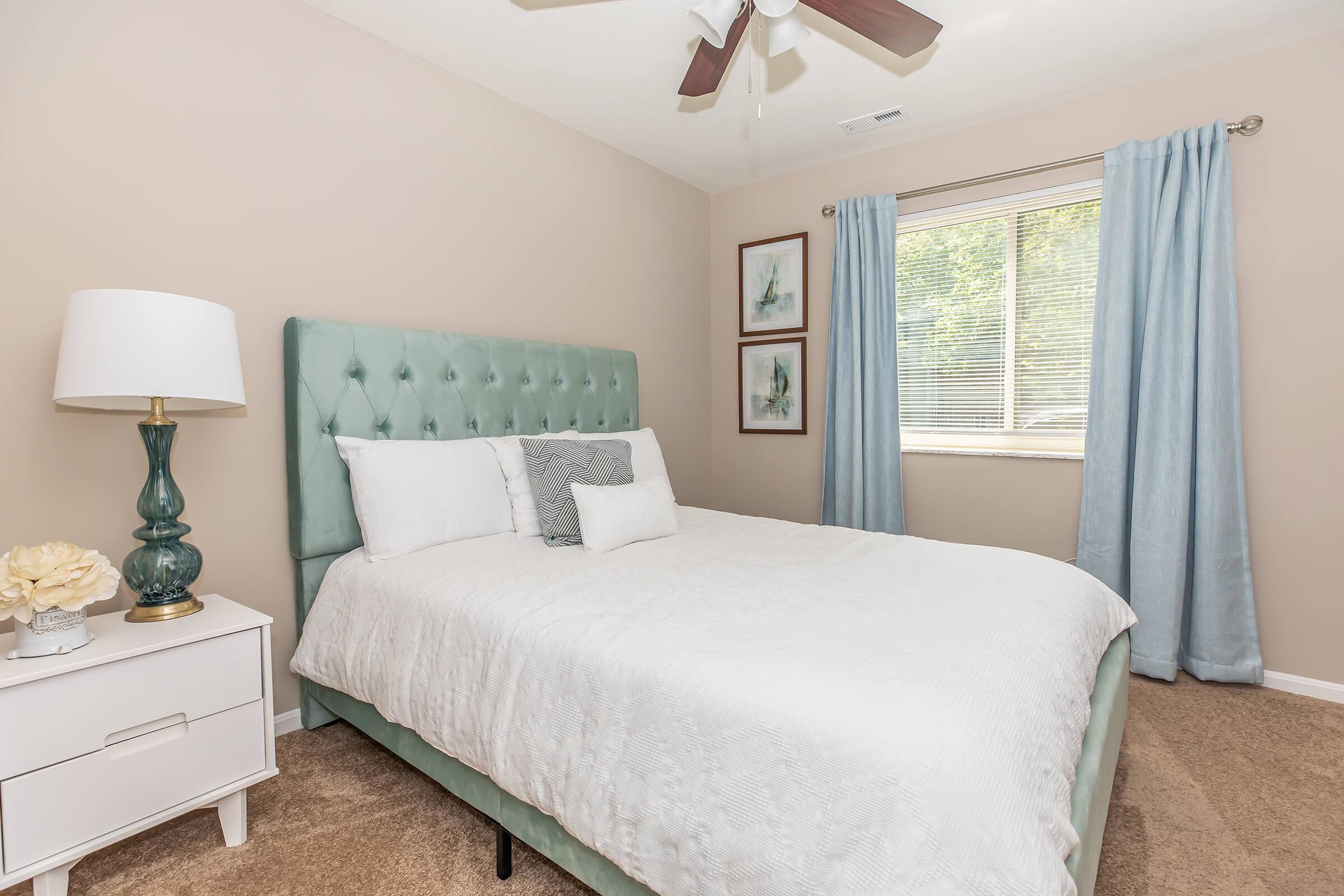 A cozy bedroom featuring a green tufted headboard, white bedding, and decorative pillows. There is a nightstand with a lamp and decorative flower, as well as two framed artworks on the wall. Large window draped with light blue curtains allows natural light to fill the space, enhancing the inviting atmosphere.