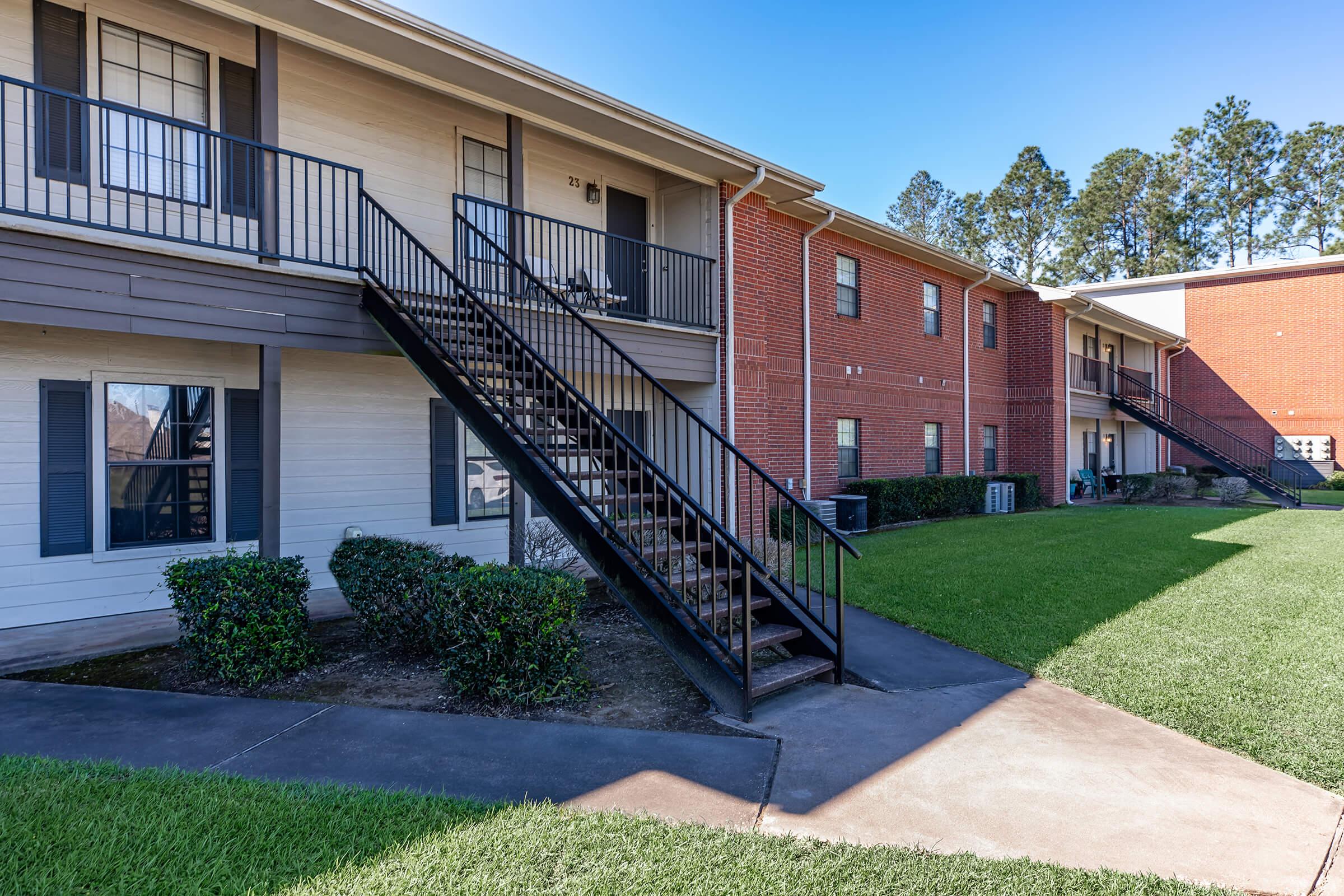 A view of a two-story apartment complex with a staircase leading to the upper floor. The buildings feature a mix of brick and siding, with landscaped grass areas in front and large windows. Clear blue sky and trees are visible in the background.