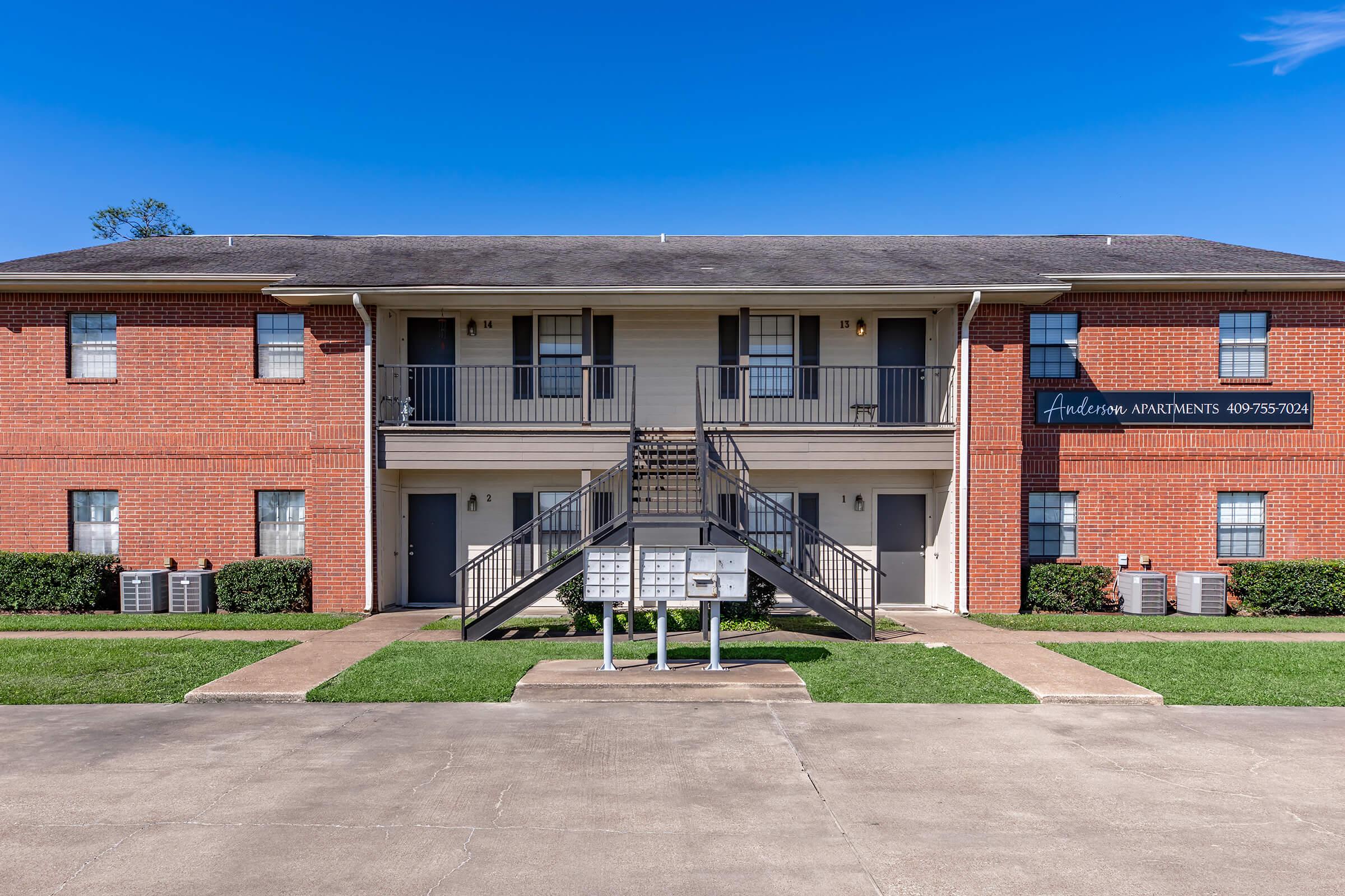 A view of a two-story apartment building with brick and white siding. Stairs lead up to the second floor, and there are two doors on each level. A row of mailboxes is positioned in front on the ground. The surrounding area features well-kept grass and a clear blue sky.