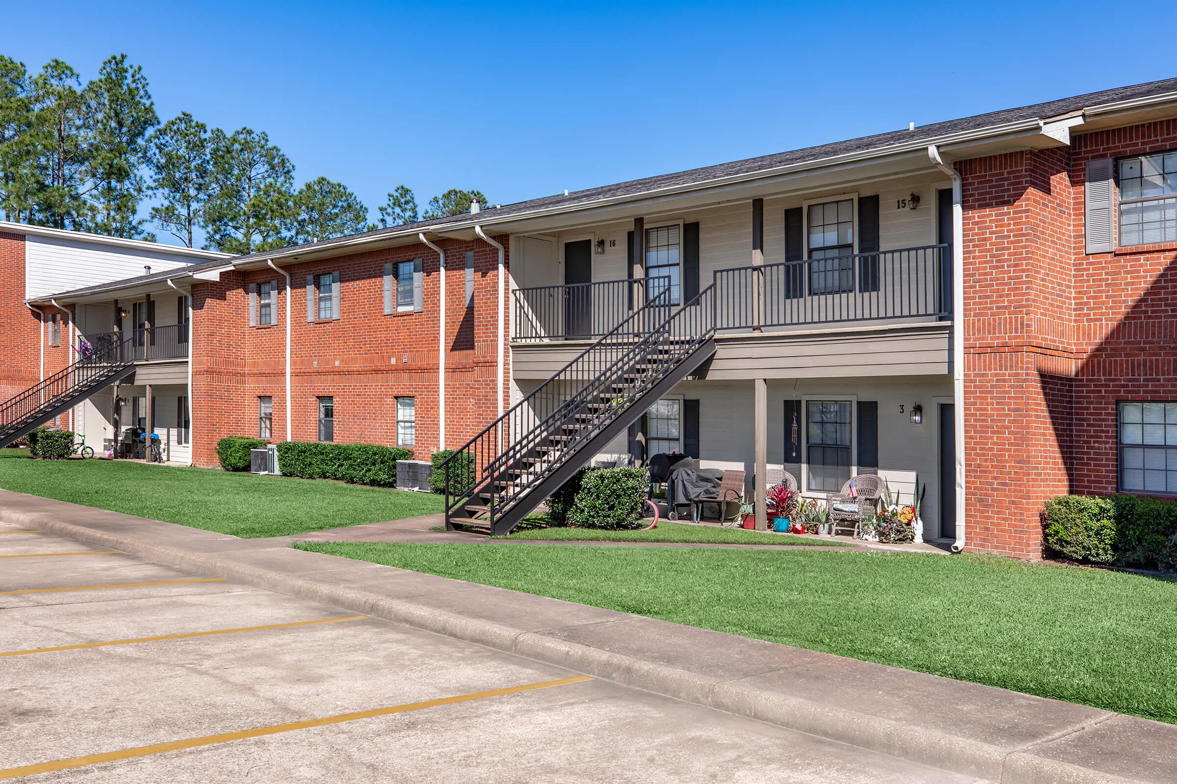 A view of a two-story apartment complex featuring brick facades and a staircase leading to upper-level units. The area is well-maintained with green grass and shrubs, and there are small sitting areas with chairs on the porches. A parking lot is visible in the foreground under clear blue skies.