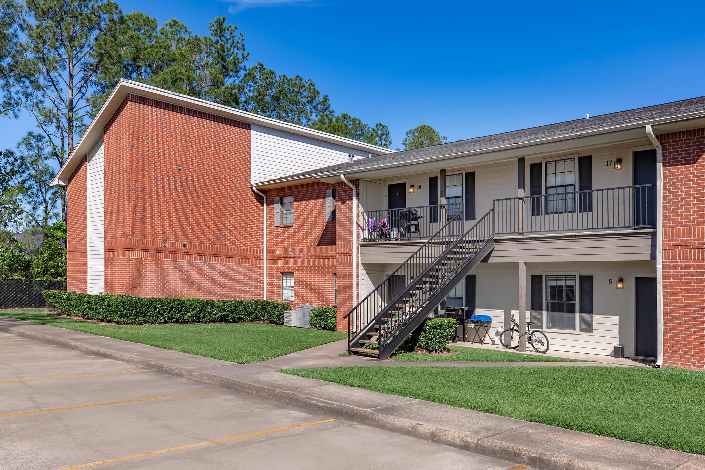 Exterior view of a residential apartment building featuring a brick and siding facade. A staircase leads to the second floor, with balconies on both levels. Two bicycles are parked near the entrance, and landscaped green grass lines the walkway. The sky is clear and sunny, creating a bright atmosphere.
