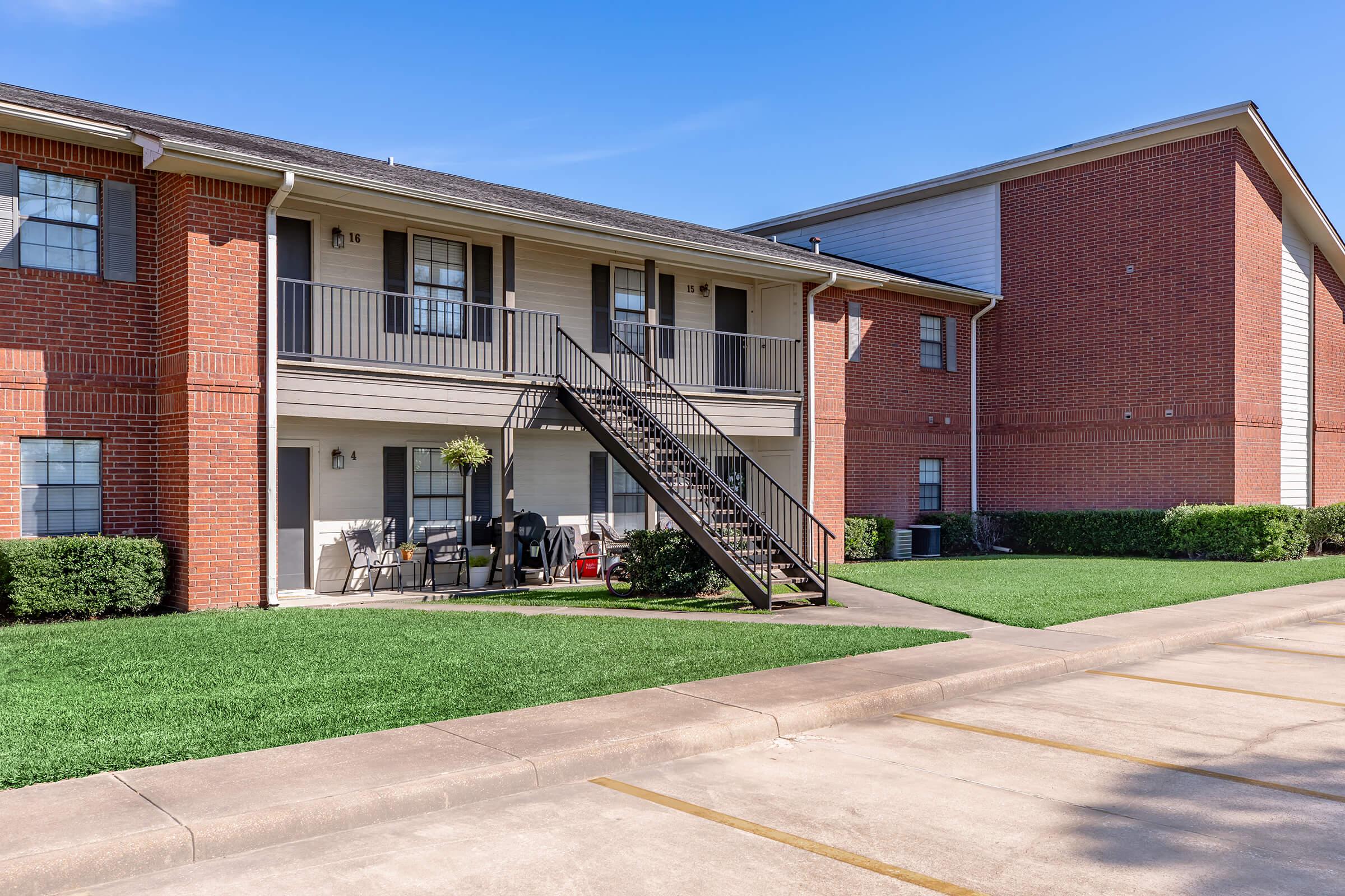 Two-story apartment building with a balcony and stairs leading up to the second floor. The exterior features brick and siding with green grass in front. A small patio area with furniture is visible. Parking spaces are shown in the foreground, with a clear blue sky overhead.
