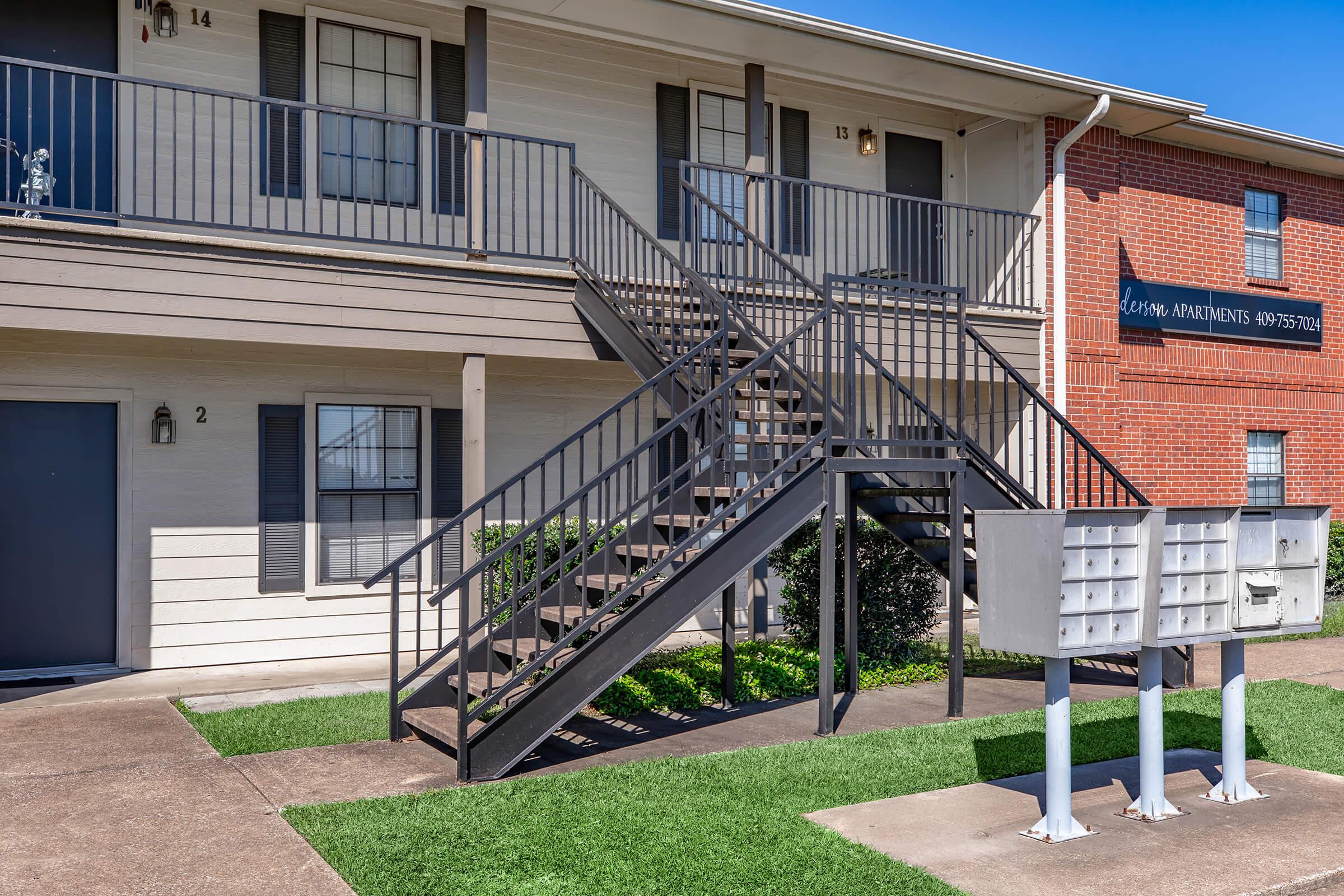 A two-story apartment building with a staircase connecting the floors. The ground level features two entrance doors, one on the left and one on the right, with a mailbox structure in front. The area is landscaped with green grass and has a clear blue sky overhead.