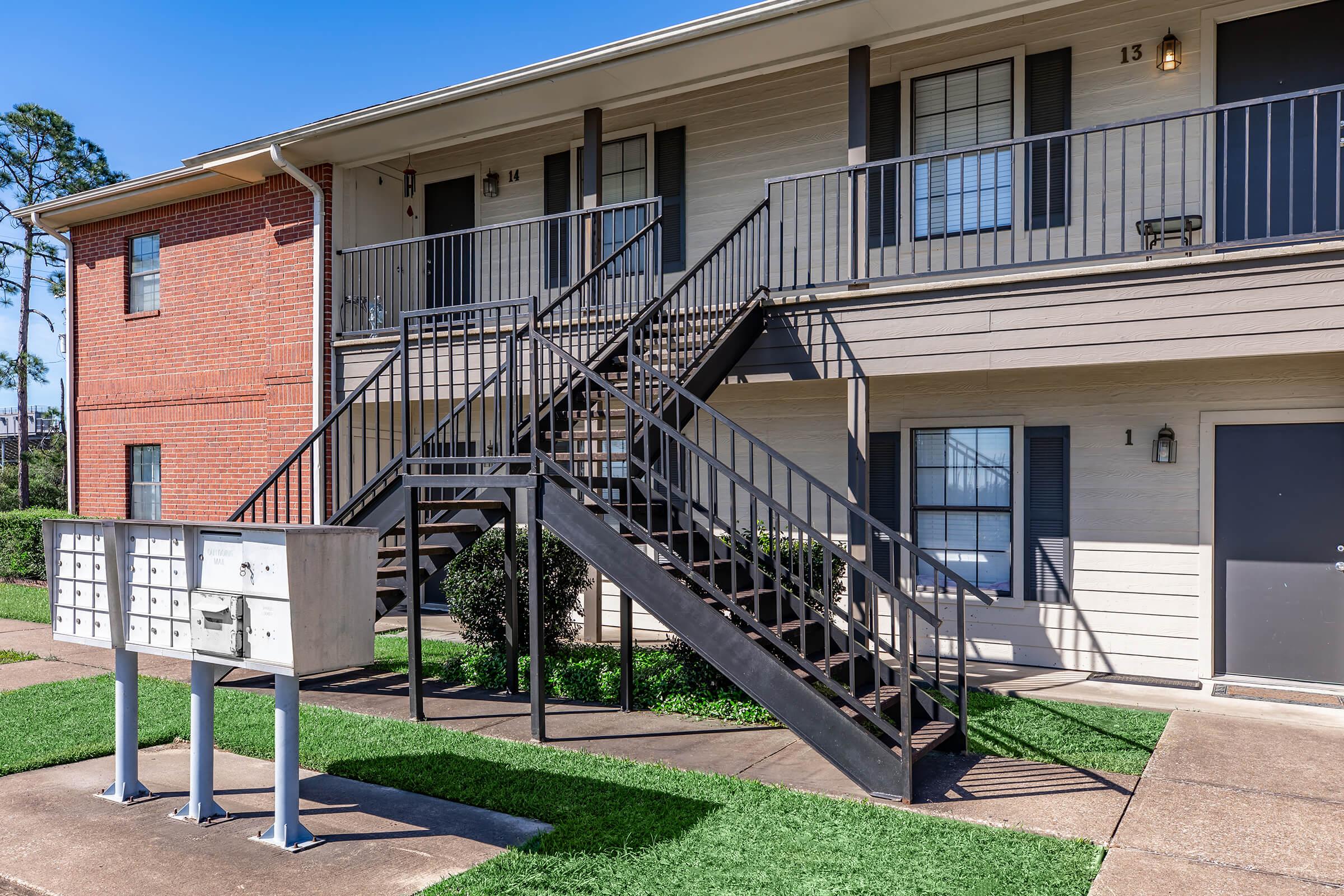 A two-story apartment building with a staircase leading to the upper floor. The ground level features mailboxes in front, and the building has a mix of brick and siding. Lush green grass lines the walkway, and a clear blue sky is visible above.