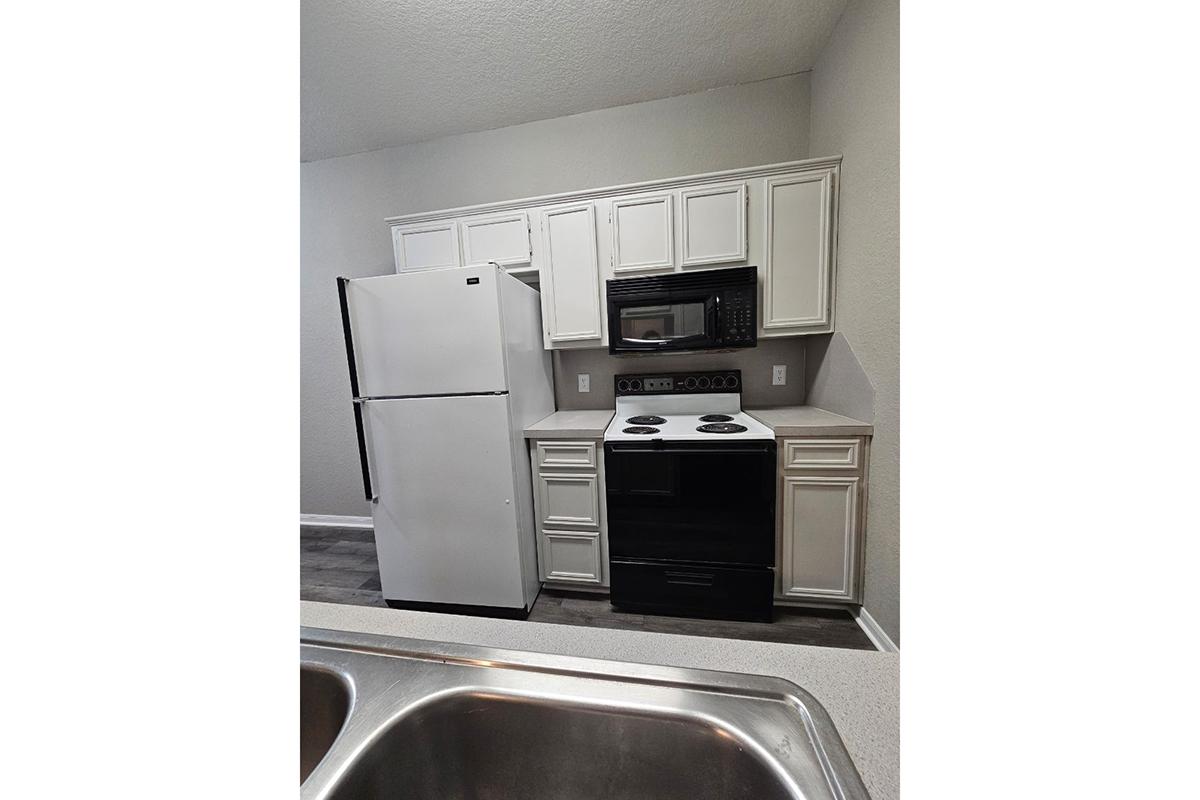 A modern kitchen featuring a white refrigerator, a black electric stove with an oven, and a microwave above it. The cabinets are white with a simple design, and the countertop is light-colored with a smooth finish. The flooring is dark, providing contrast to the cabinetry.