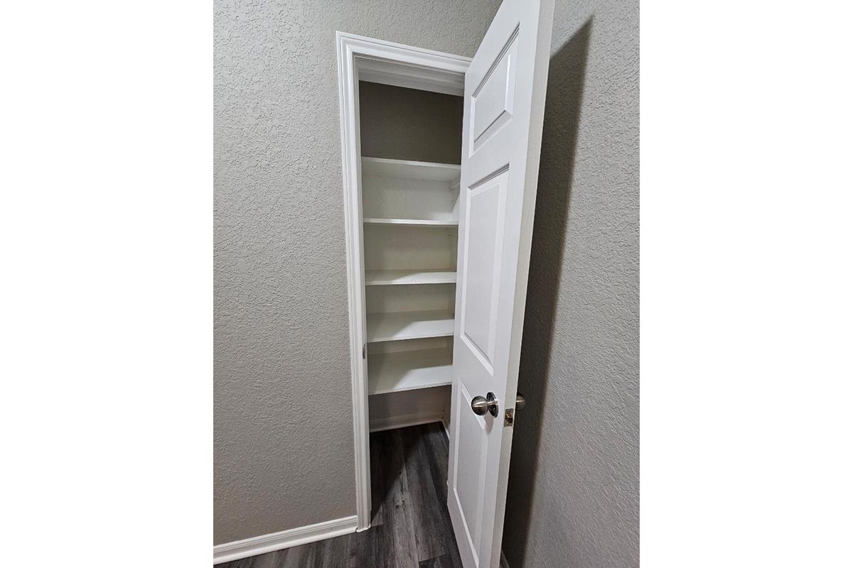 A white door partially open, revealing a small pantry or storage closet with five white shelves against a light gray wall. The floor is a dark wood laminate, giving a modern appearance to the space.
