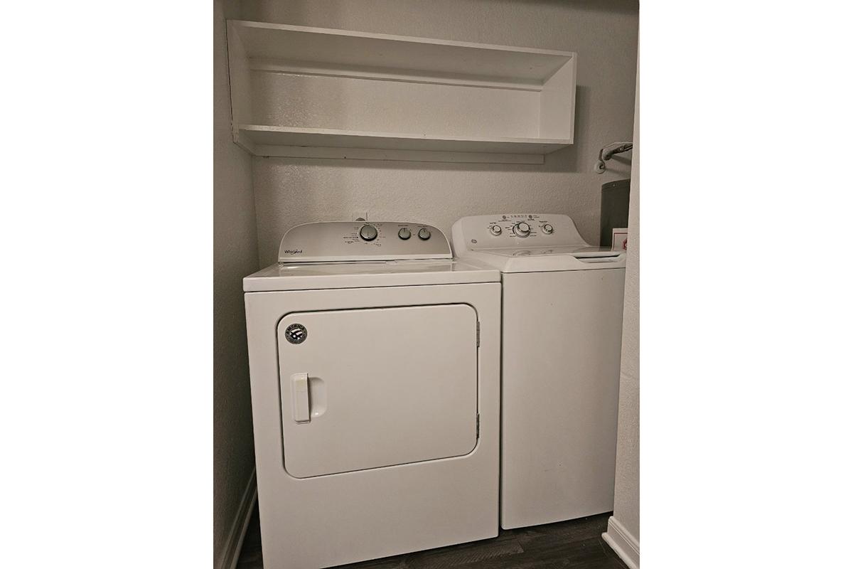 A stacked white washer and dryer set in a laundry room with a shelf above for storage. The appliances have controls on the front, and the space features neutral-colored walls and flooring.