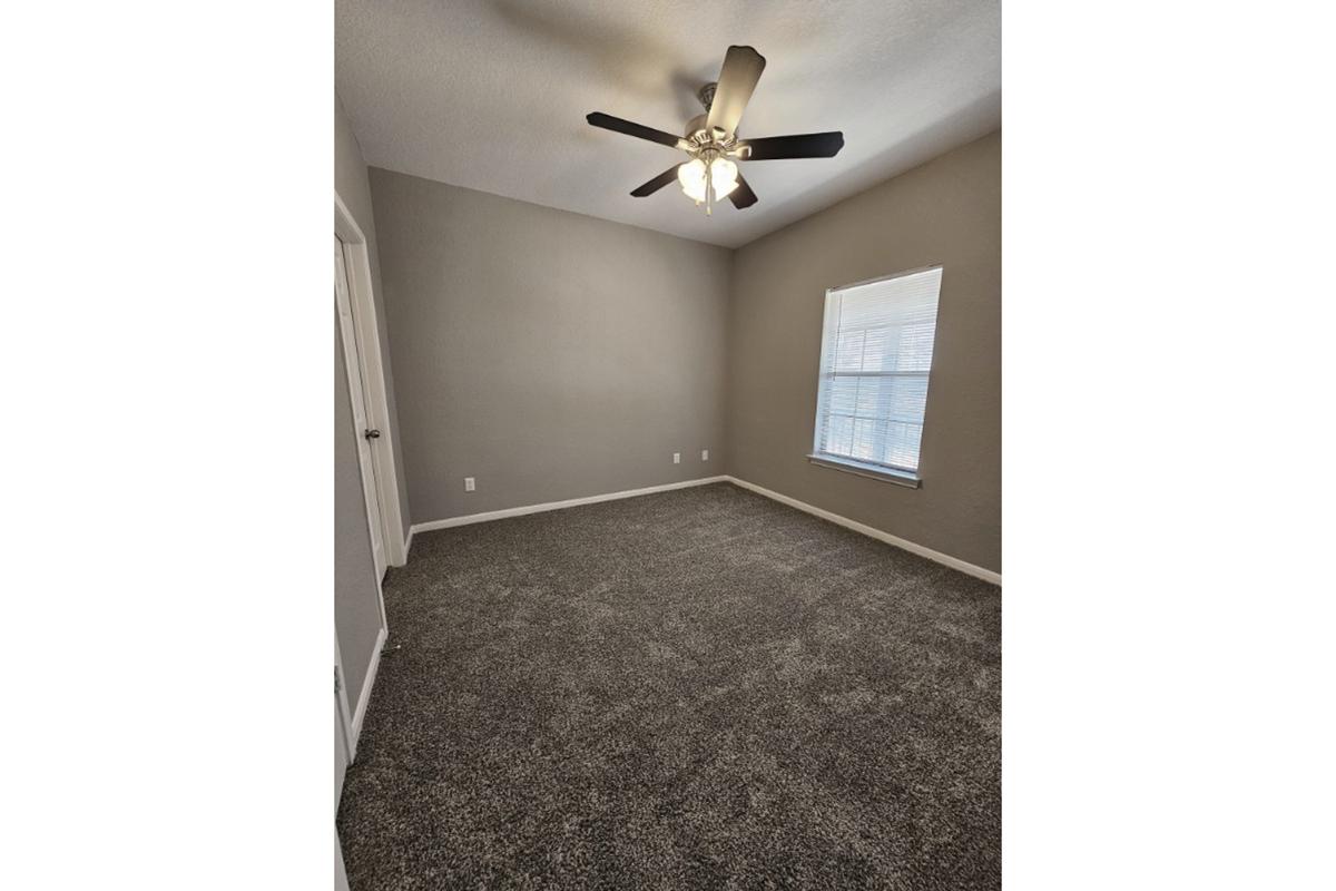 A vacant room with light gray walls and a textured carpet. The ceiling features a ceiling fan with light fixtures. A window allows natural light, and there is a door on one side of the room. The space is tidy and minimalist, suitable for various furniture arrangements.
