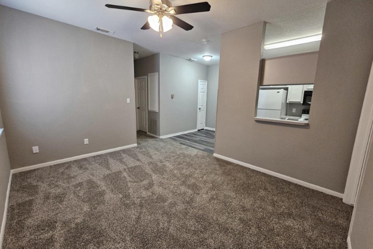 A spacious, empty living room with light gray walls and plush carpet flooring. It features a ceiling fan, a view into a hallway leading to a door, and an open kitchen area with white appliances visible through a cutout. Natural light brightens the space.