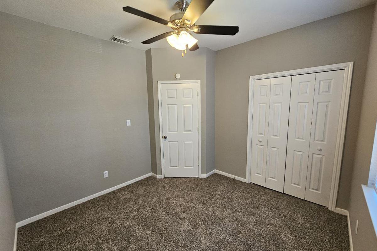 A small, well-lit room featuring light gray walls and carpet. It includes a ceiling fan with light, a single door leading to the hallway, and double closet doors. A window allows natural light to enter, providing a cozy atmosphere. The room is empty, ready for furnishings.