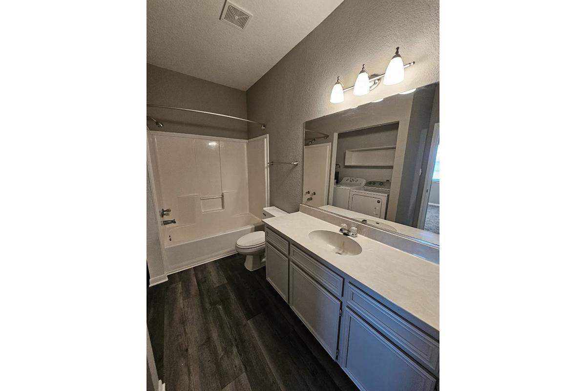 A clean and modern bathroom featuring a white bathtub and shower combo, a double sink vanity with light gray cabinetry, a toilet, and dark vinyl flooring. The room is well-lit with three overhead light fixtures and has a neutral color palette. A laundry area is visible in the background.