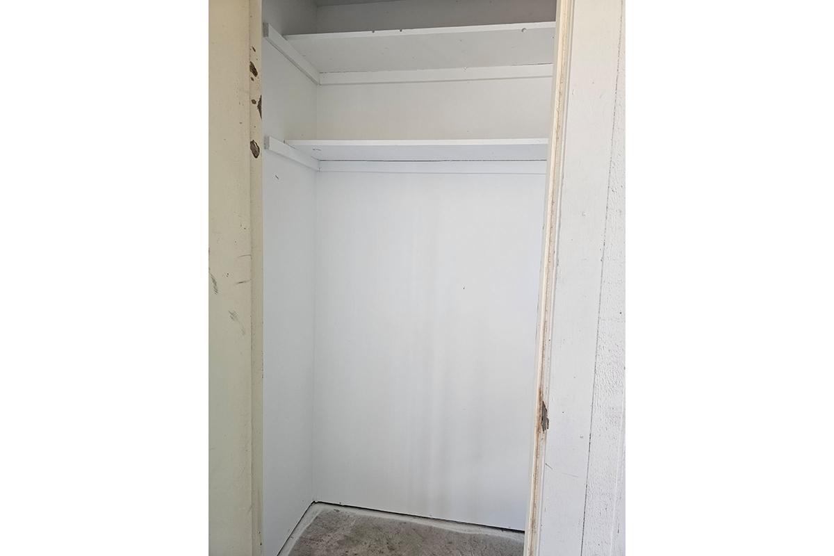 An empty closet with plain white walls and two shelves. The floor appears bare, and the interior is well-lit, providing a clean, minimalist appearance. The closet door is partially open, revealing the lack of any stored items inside.
