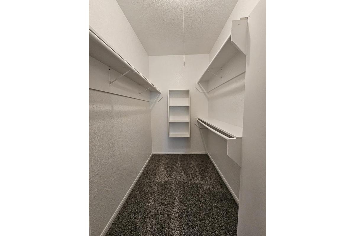 A spacious, empty closet featuring white walls, a textured floor, and several mounted shelves on either side. In the center, there is a narrow shelving unit against the back wall, providing additional storage space. The overall design is minimalist and bright, creating an organized appearance.