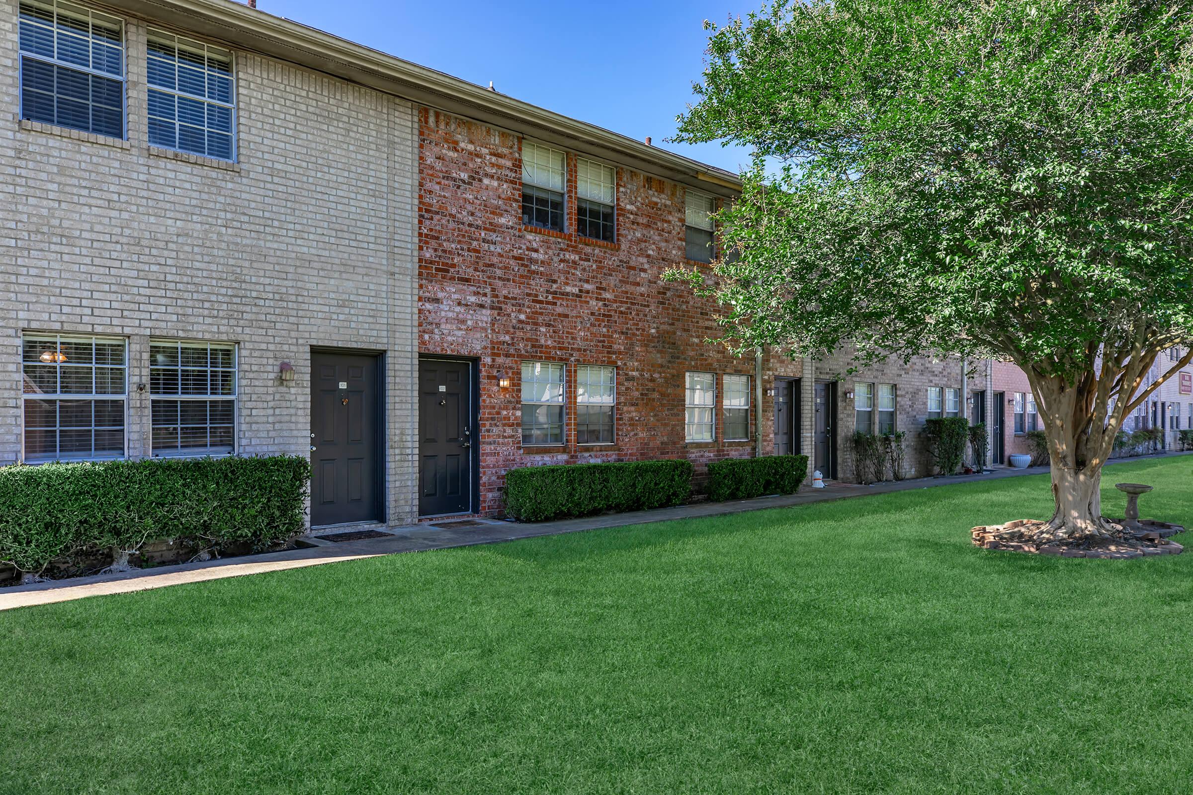 A row of two-story residential buildings featuring a mix of brick and siding exteriors. Each unit has a front door, with windows spaced evenly. A well-maintained green lawn and bushes line the walkway, complemented by small trees providing shade. The sky is clear and blue, suggesting a sunny day.