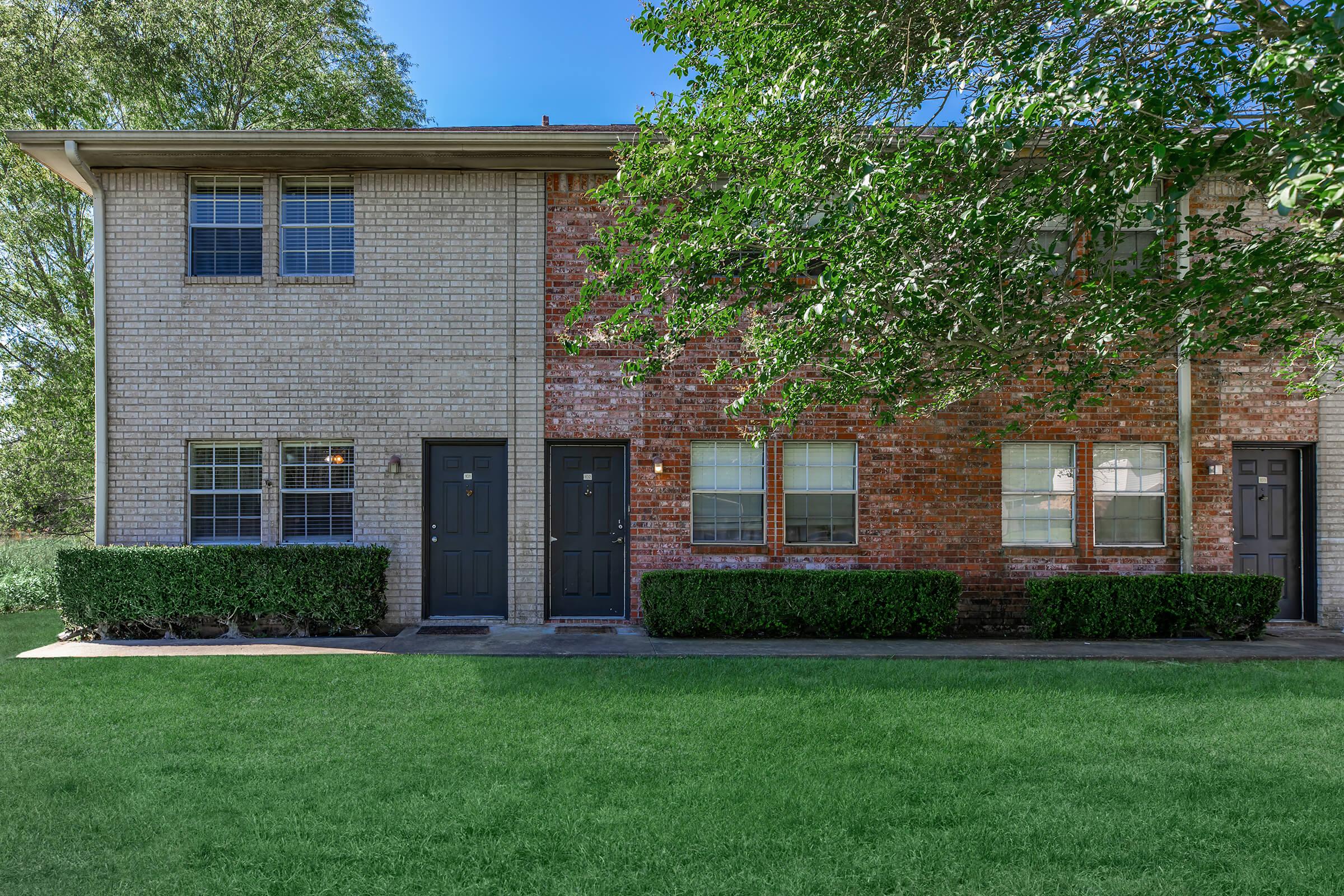 A two-story brick apartment building with a mix of red and tan brick exterior. The front features two identical doors with dark frames, large windows, and a well-manicured lawn with green grass. Trees provide shade on one side, and the sky is clear and blue in the background.