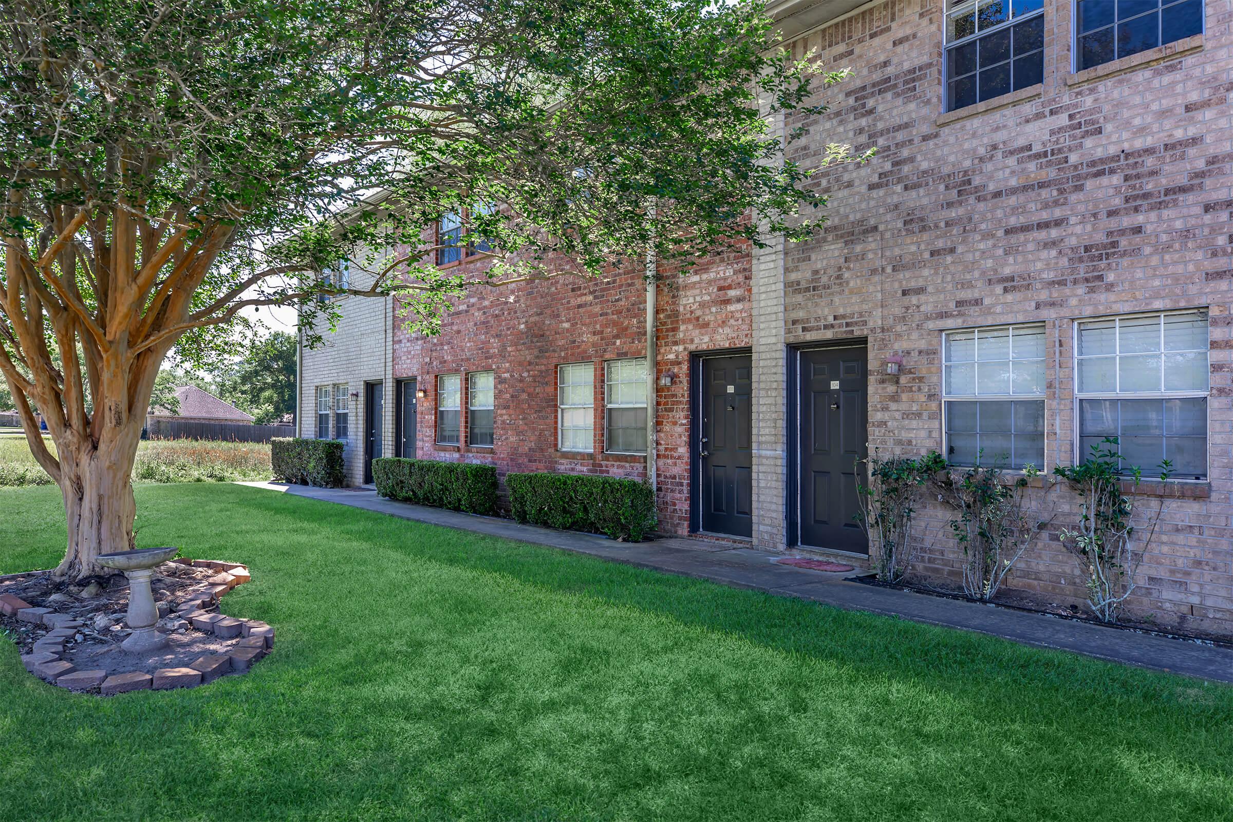 A row of brick apartment buildings with two stories, featuring green lawns and neatly trimmed bushes. A small tree with a decorative base is positioned in front of the buildings, and the scene is bathed in natural sunlight. The atmosphere is peaceful and well-maintained.