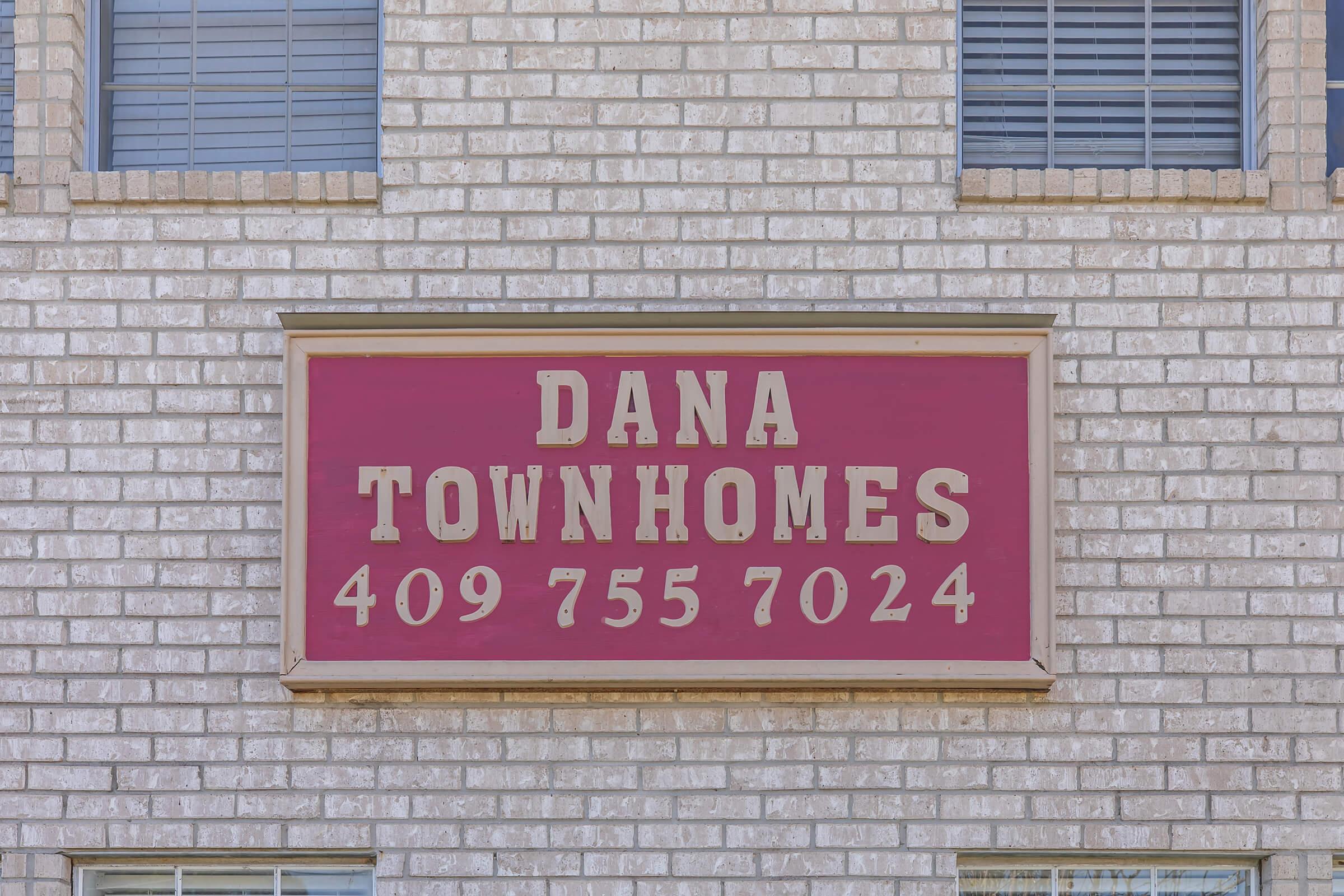 Sign for Dana Townhomes displayed on a brick wall, featuring the name "DANA TOWNHOMES" in large white letters against a maroon background, along with the phone number "409 755 7024" underneath. Windows with blinds are visible above the sign.