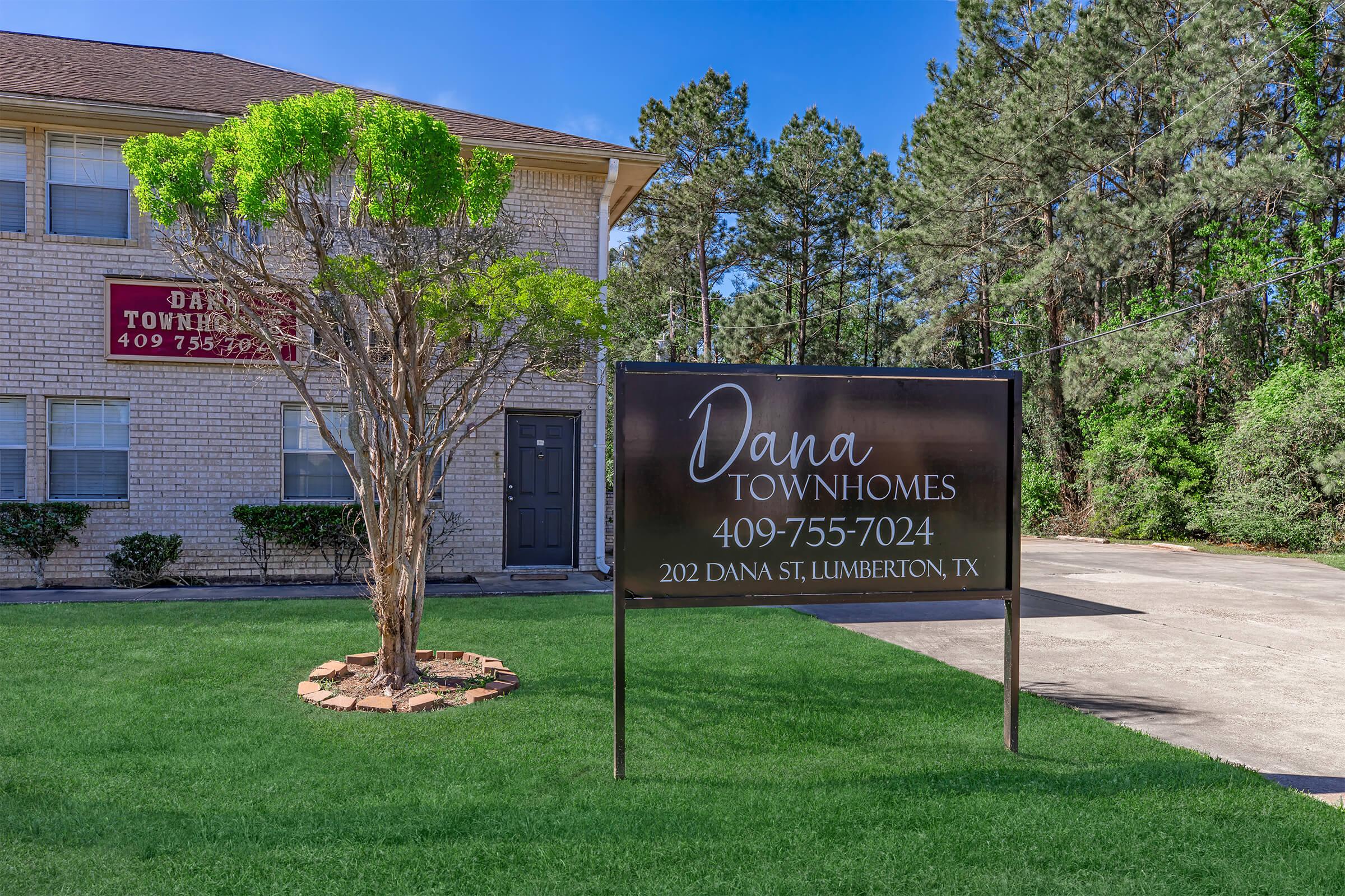 Sign for Dana Townhomes located at 202 Dana St, Lumberton, TX, displaying the phone number 409-755-7024. The background features green grass, a tree, and a multi-story building surrounded by trees.