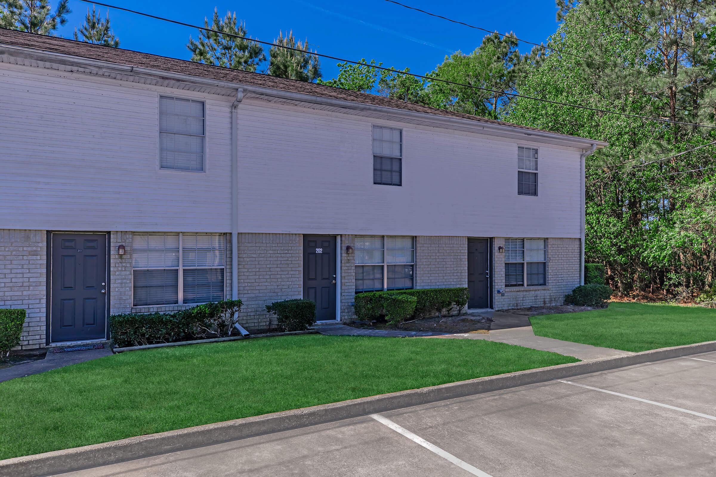 A row of two-story residential buildings with white siding and gray doors. Each unit has a small front porch, surrounded by manicured lawns and shrubs. The scene includes a clear blue sky and trees in the background, with a paved parking area in the foreground.