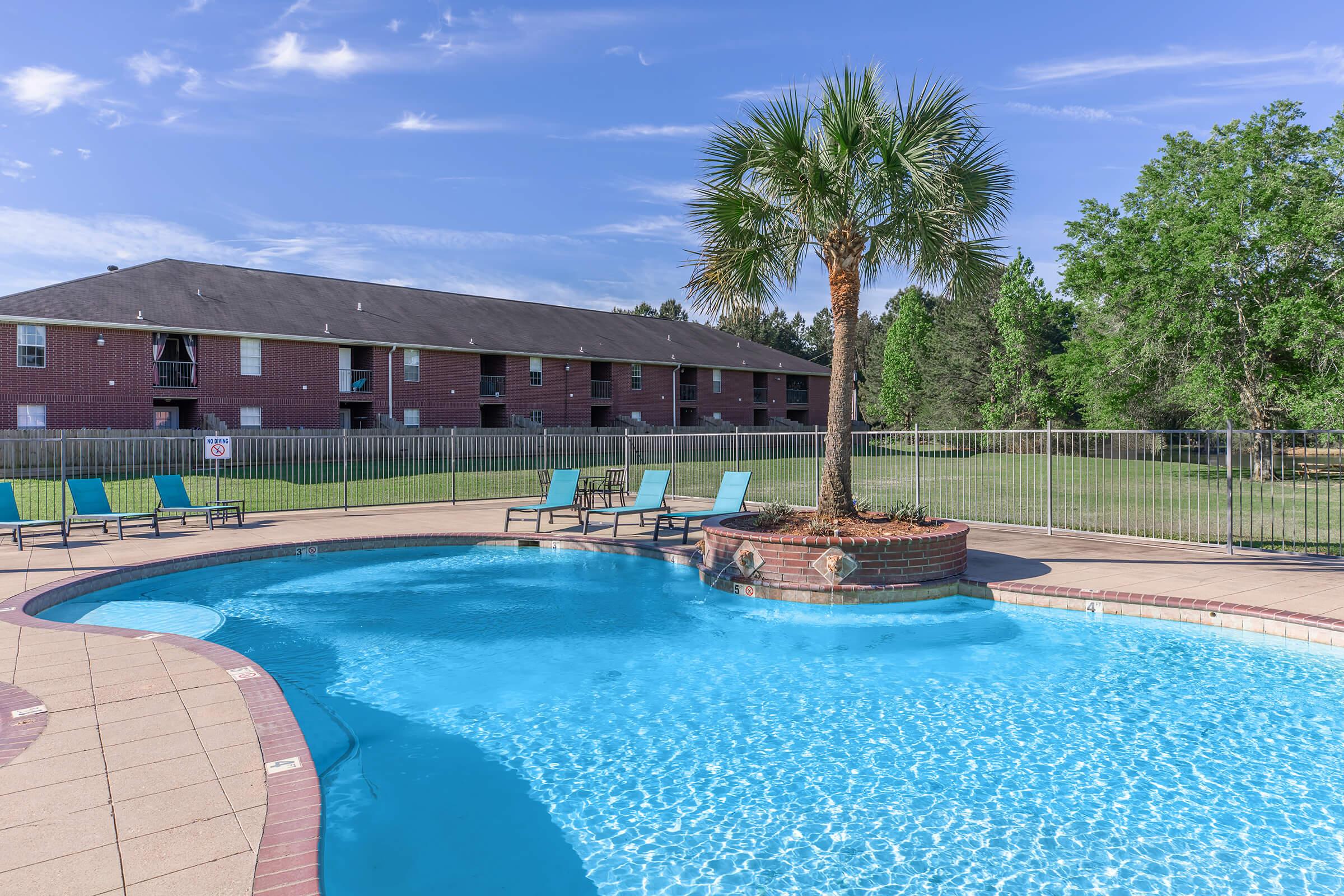 A clear swimming pool surrounded by a tiled patio, equipped with lounge chairs in shades of blue and yellow. In the center of the pool, there is a small landscaped area with a palm tree. In the background, a two-story apartment building and green trees can be seen under a blue sky with light clouds.