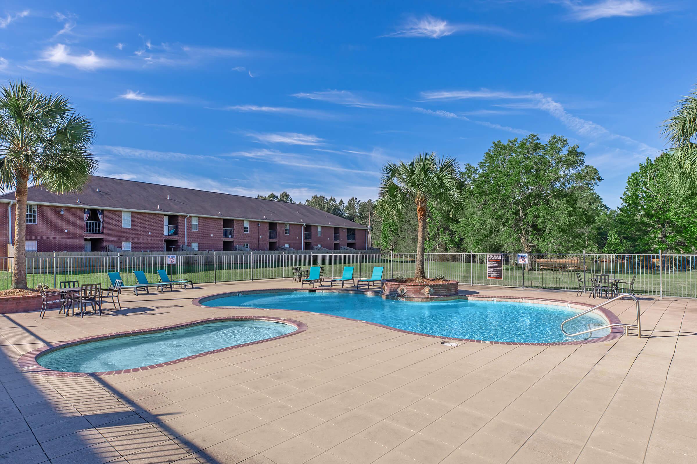 A sunny outdoor pool area featuring a large swimming pool with a connected hot tub. Surrounding the pool are lounge chairs in vibrant colors and small tables. Lush green trees and a grassy area are visible in the background, along with a multi-unit residential building. Clear blue skies add to the inviting atmosphere.