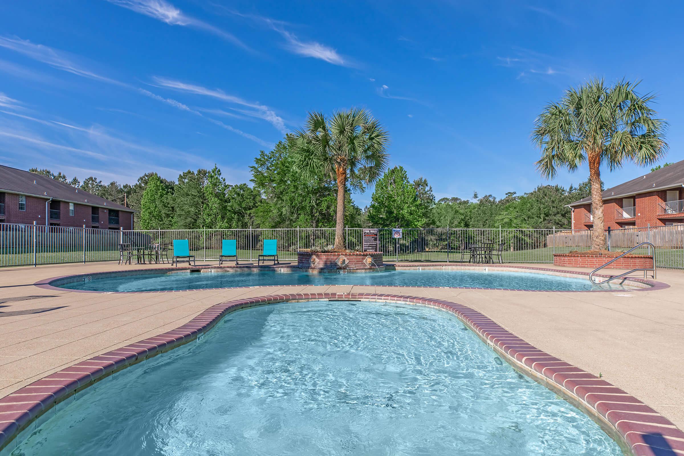 View of a serene swimming pool area featuring a main pool with blue water, a separate section for relaxation, lounge chairs in vibrant colors, and lush greenery in the background. Palm trees add a tropical feel, and the sky is clear with scattered clouds, creating a peaceful outdoor atmosphere.