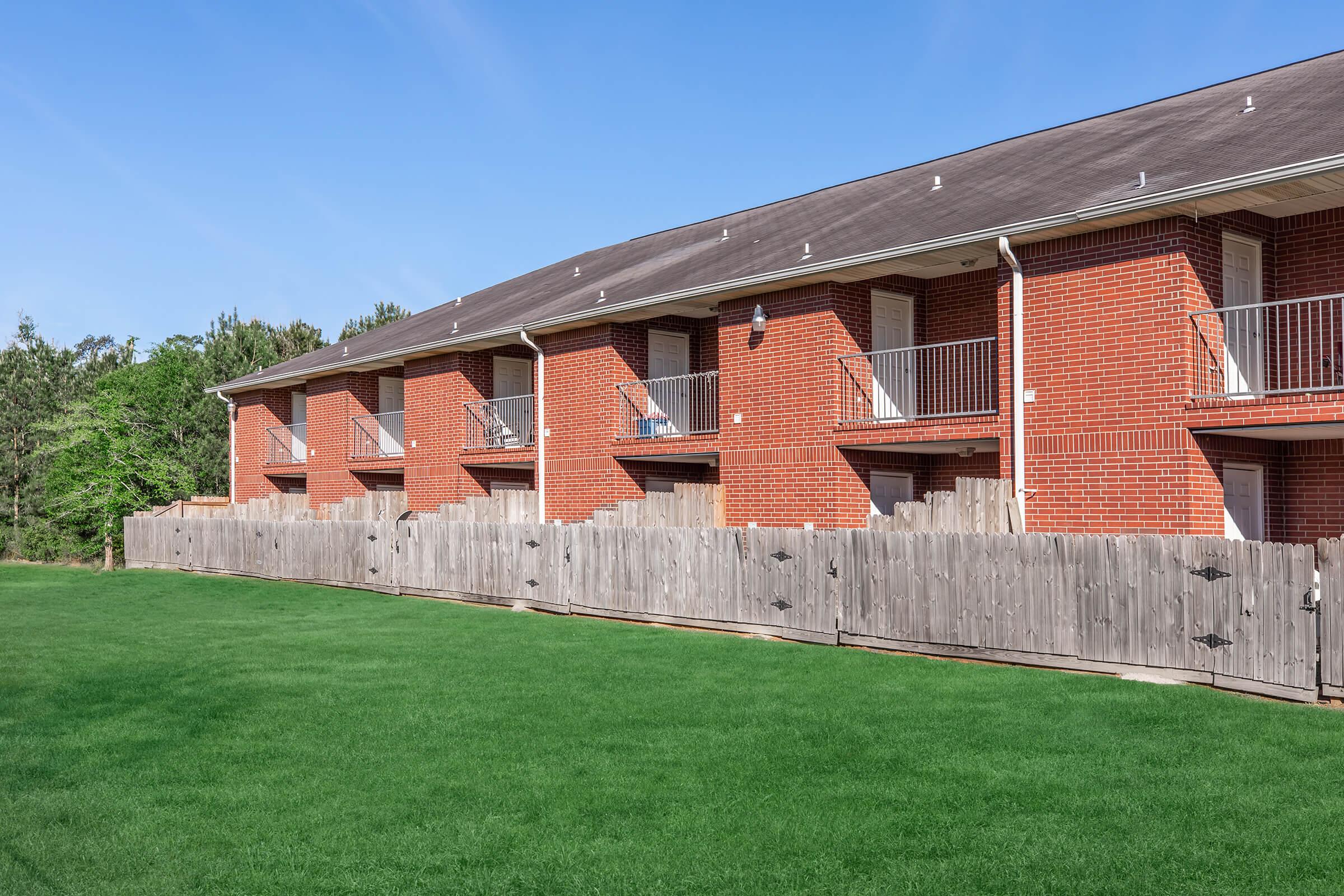 A two-story apartment building made of red brick, featuring multiple balconies. There is a neatly maintained green lawn in front, bordered by a wooden fence with two gates. The sky is clear and blue, with a few trees visible in the background.