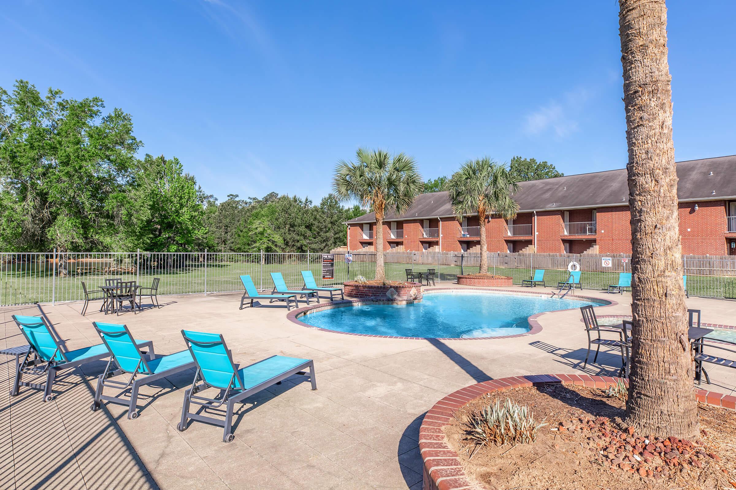 A serene outdoor pool area featuring a round pool surrounded by lounge chairs. There are palm trees nearby, and a brick pathway leads to the pool. In the background, a two-story brick building is visible, set against a clear blue sky and lush green trees.