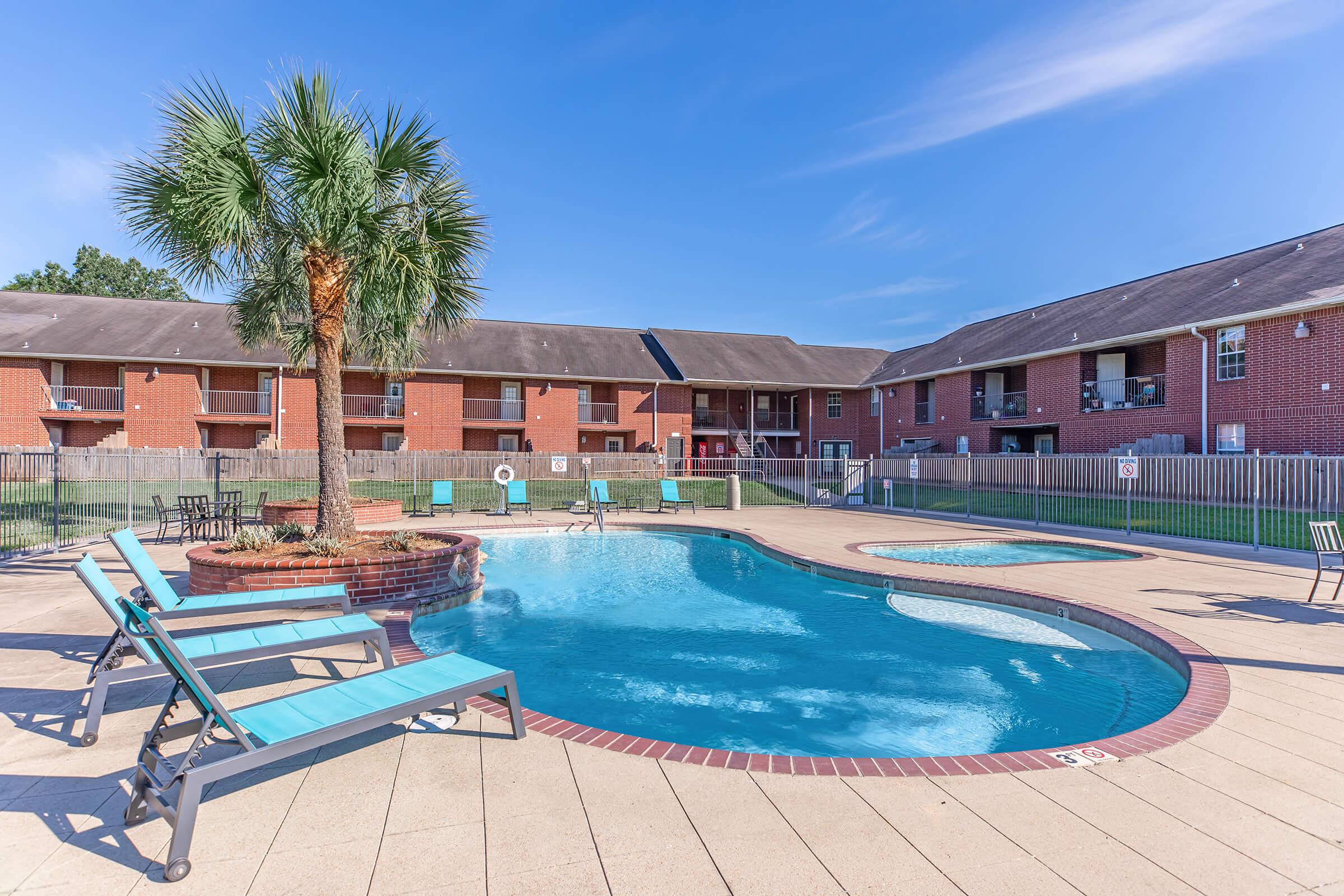 Swimming pool area with a palm tree, lounge chairs, and hot tub. Surrounding buildings are red brick apartments, with green grass and a clear blue sky above. The pool features a curved shape and is enclosed by a fence, creating a relaxing outdoor atmosphere.