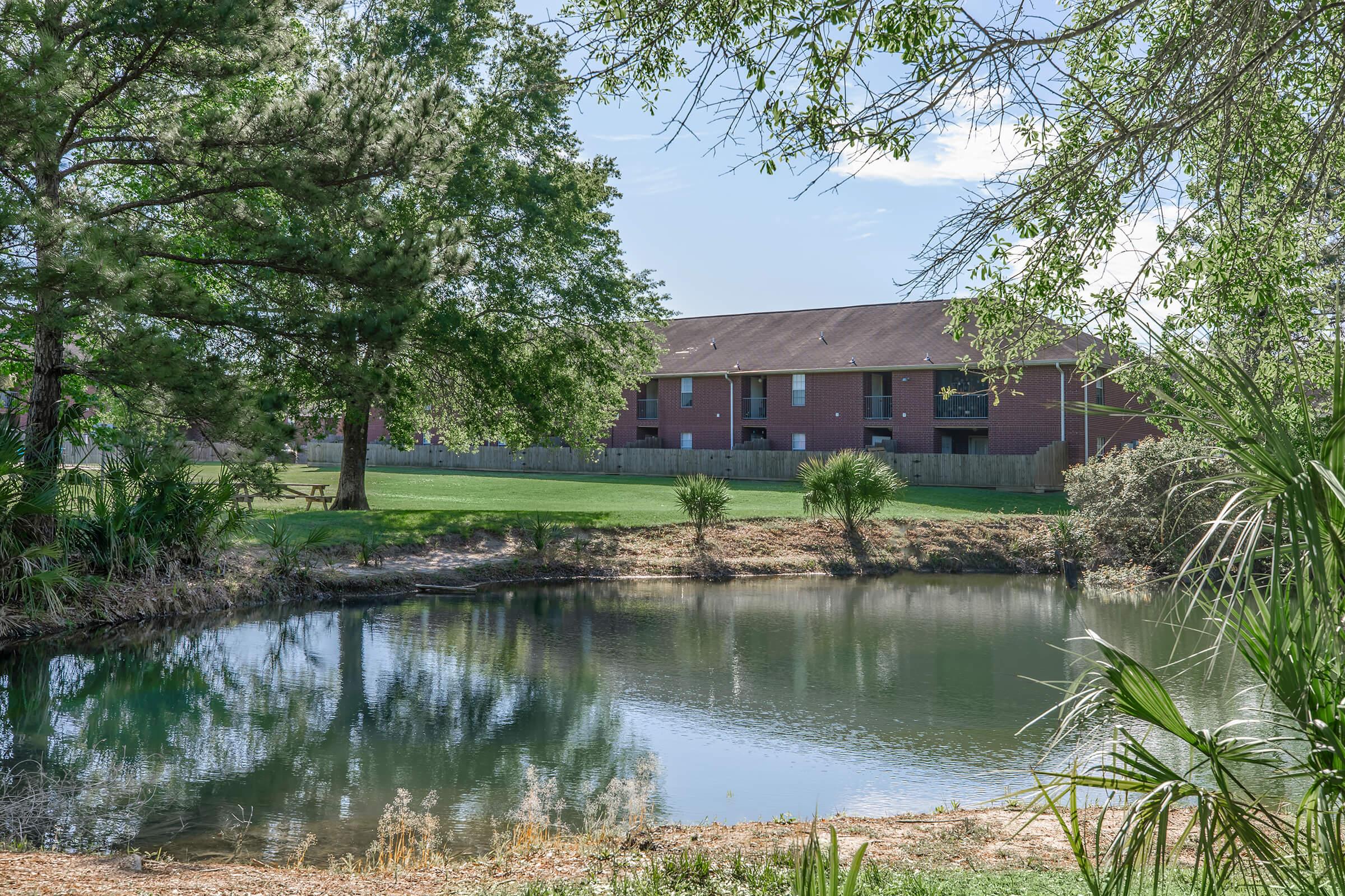 A serene pond surrounded by lush greenery, with a backdrop of a two-story brick apartment building. Trees and bushes frame the scene, creating a peaceful outdoor environment. The sky is clear with a few clouds, adding to the tranquil atmosphere.