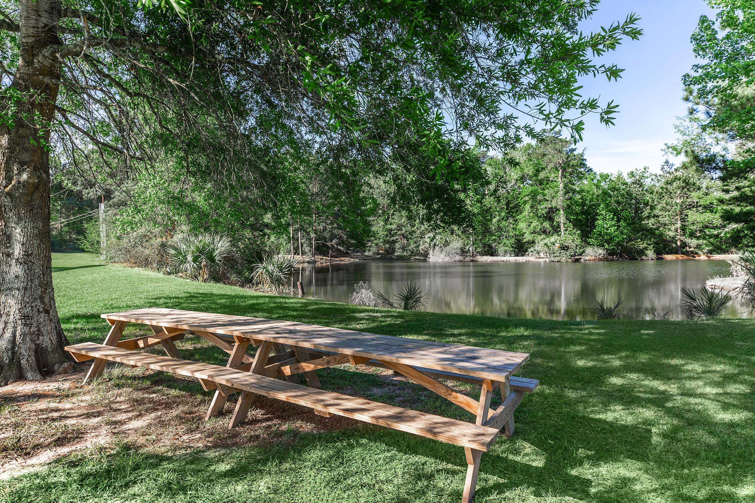 A serene outdoor scene featuring a wooden picnic table next to a calm pond. Surrounding greenery includes trees and plants, creating a peaceful atmosphere. The sky is clear, adding to the tranquility of the setting, ideal for relaxation or gatherings.