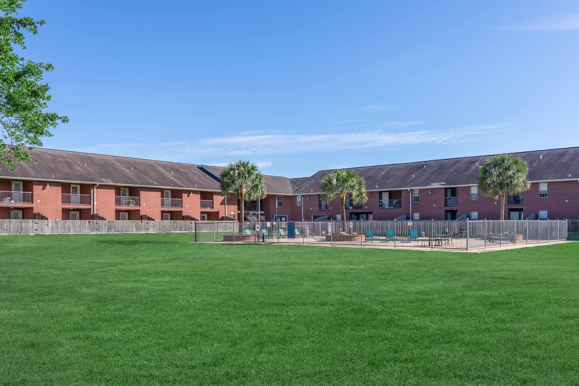 View of a residential complex featuring two buildings with red brick exteriors. The grassy area in front includes a fenced playground with benches and palm trees, under a clear blue sky. The setting conveys a peaceful suburban atmosphere.
