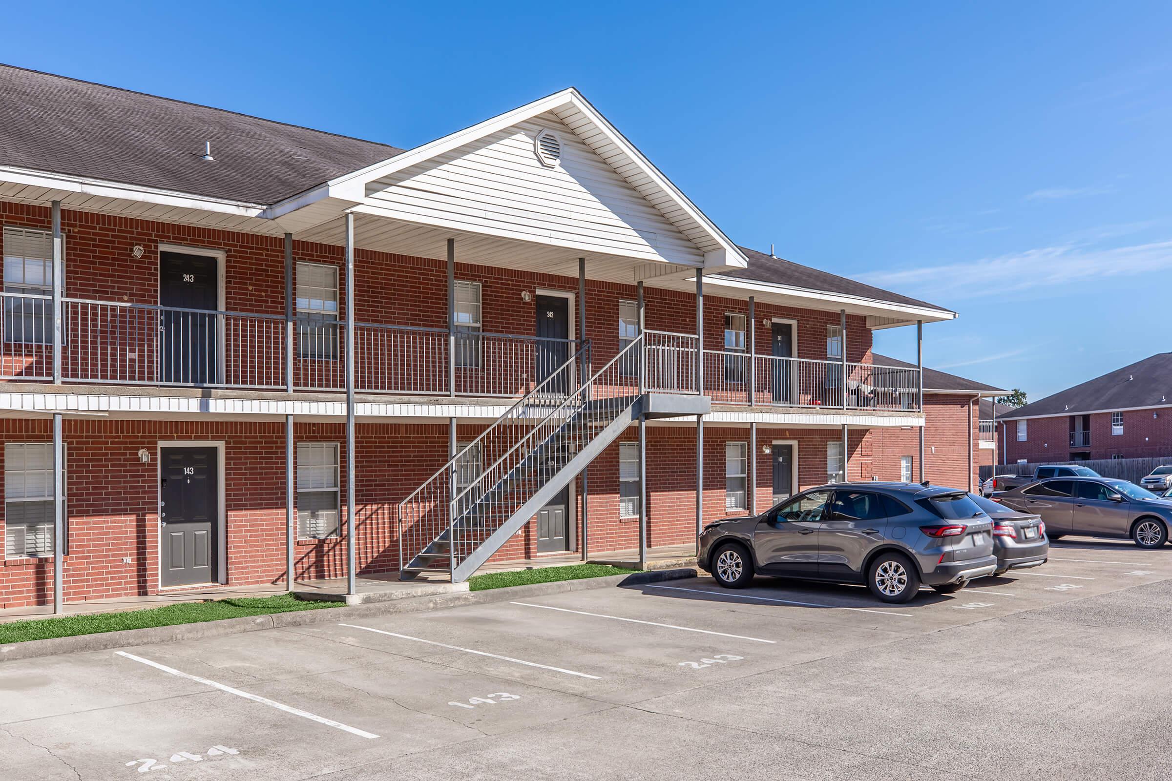 Exterior view of a two-story brick apartment building with a staircase leading to the second floor. Several parking spaces are visible in front, with one car parked. The sky is clear and blue, and the building has multiple doors on each level.